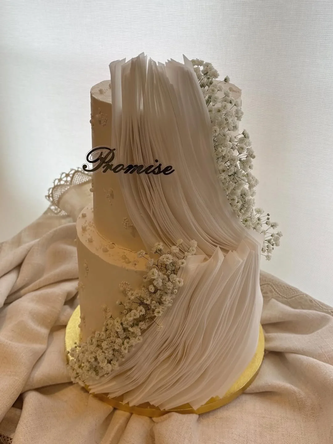 A white wedding cake decorated with baby's breath flowers and white paper or fabric resembling an open book, with a 'Promise' topper, on a gold-colored base on beige fabric.