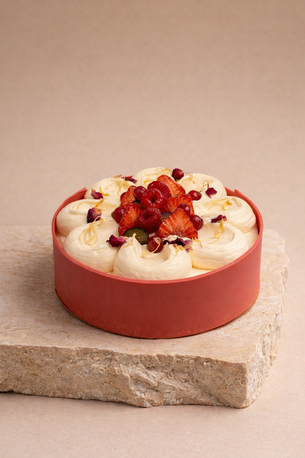 Round cake with white frosting decorated with fresh strawberries, red berries, and edible flower petals, placed on a stone slab.