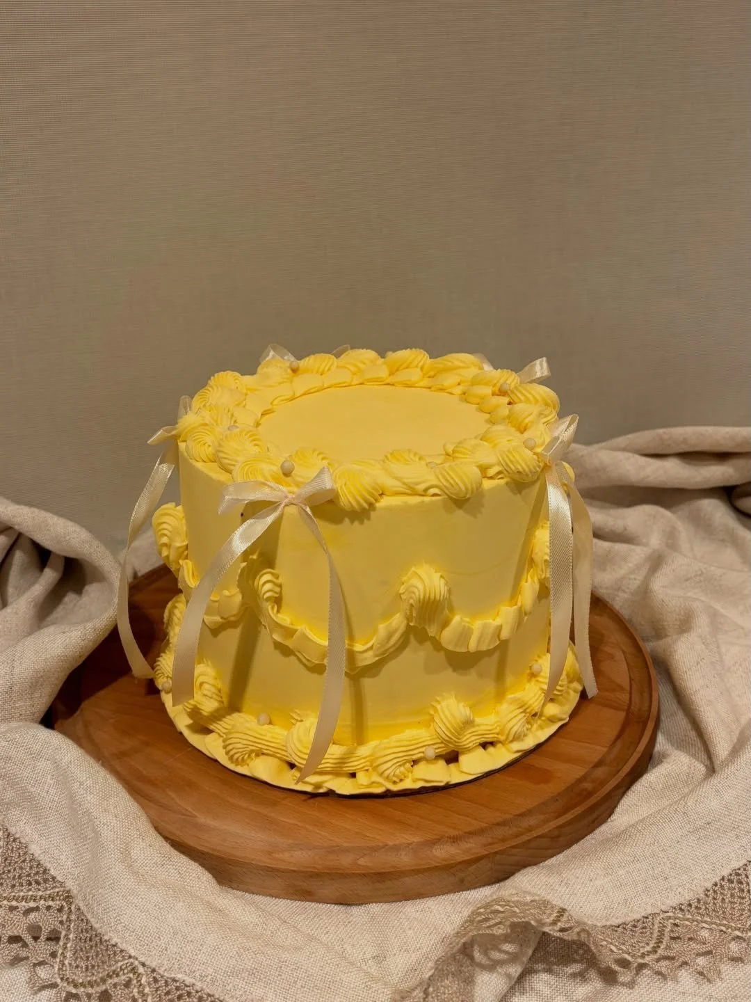 Yellow layered cake with piped cream decorations, bows, and small pearls, placed on a wooden board with a fabric backdrop.