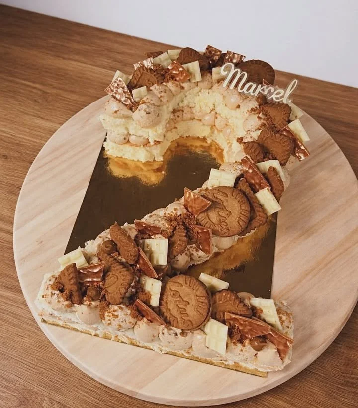Number 0 shaped cake decorated with chocolate, white chocolate, and cookies, topped with a cake topper that spells 'Maxel'.