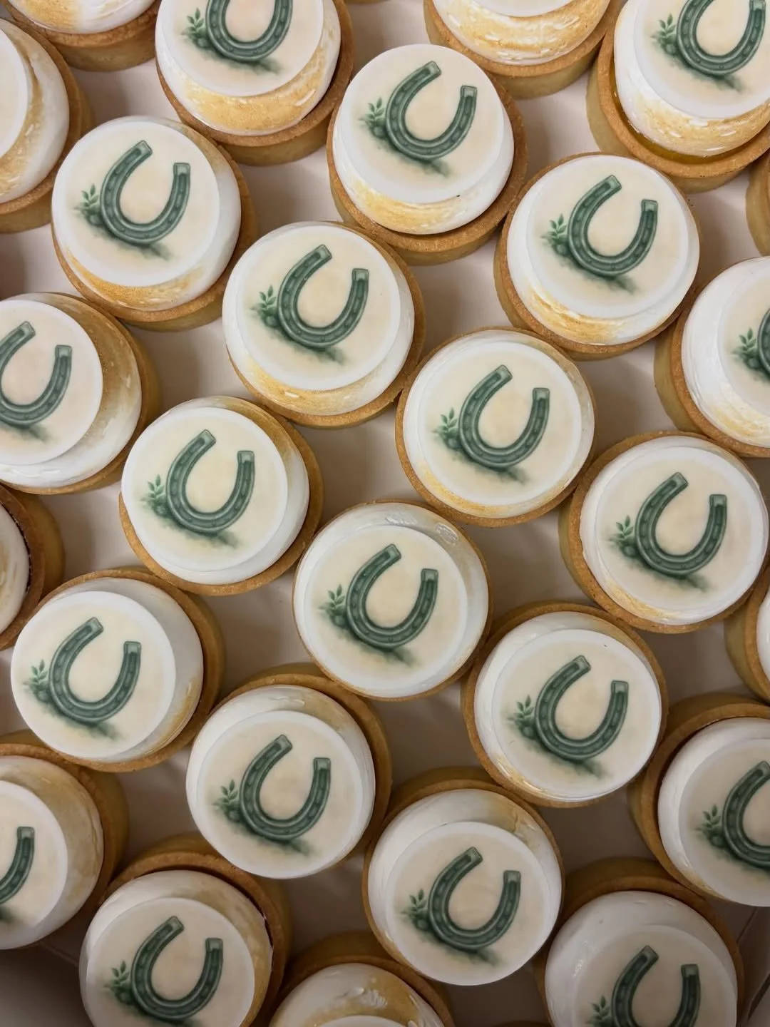 Multiple small cupcakes with white frosting and a green horseshoe decoration on top.