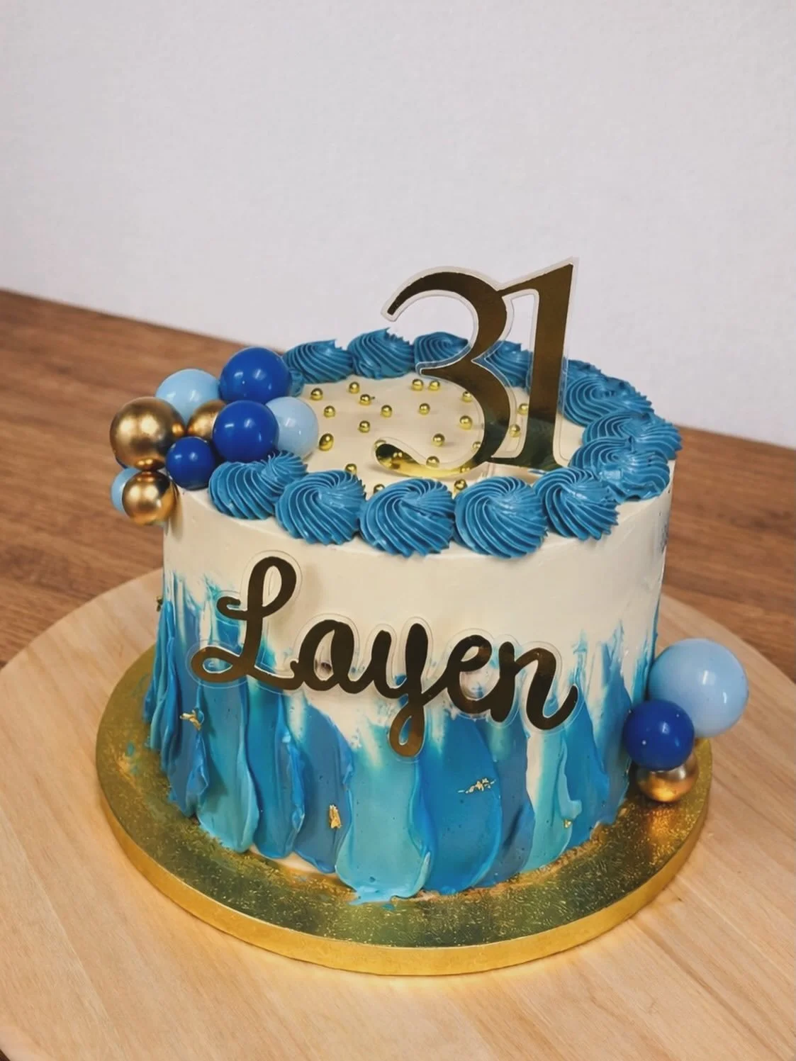Celebration cake with blue and white marbled icing, blue frosting rosettes around the top edge, decorative gold and blue balloons on the side, and a black 'Lauen' topper with a large gold '31' on top, indicating a 31st birthday.