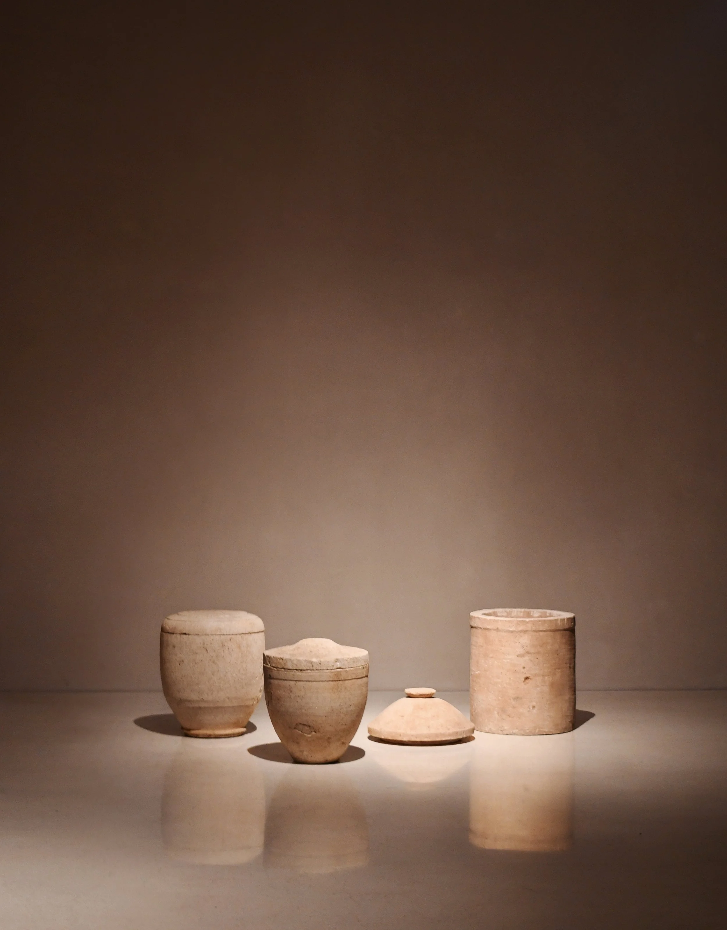 Seven ancient clay pots of various sizes and shapes are displayed against a plain, dark background. The pots are light-colored and appear to be handmade, with some showing signs of age and wear.