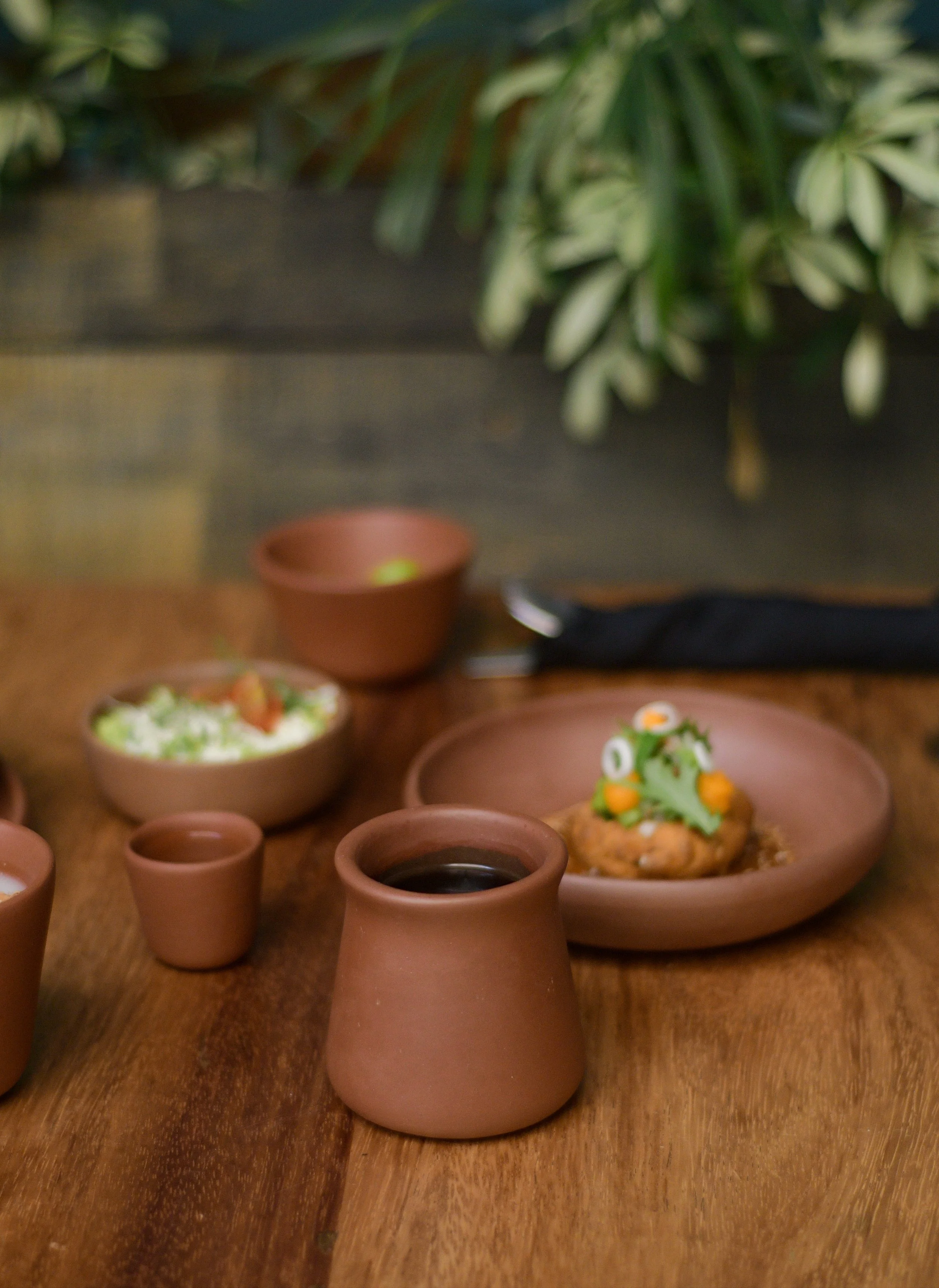 A blurry image of a dining table with multiple terracotta cups and bowls, some filled with food and drink, and a plant with variegated leaves in the background.