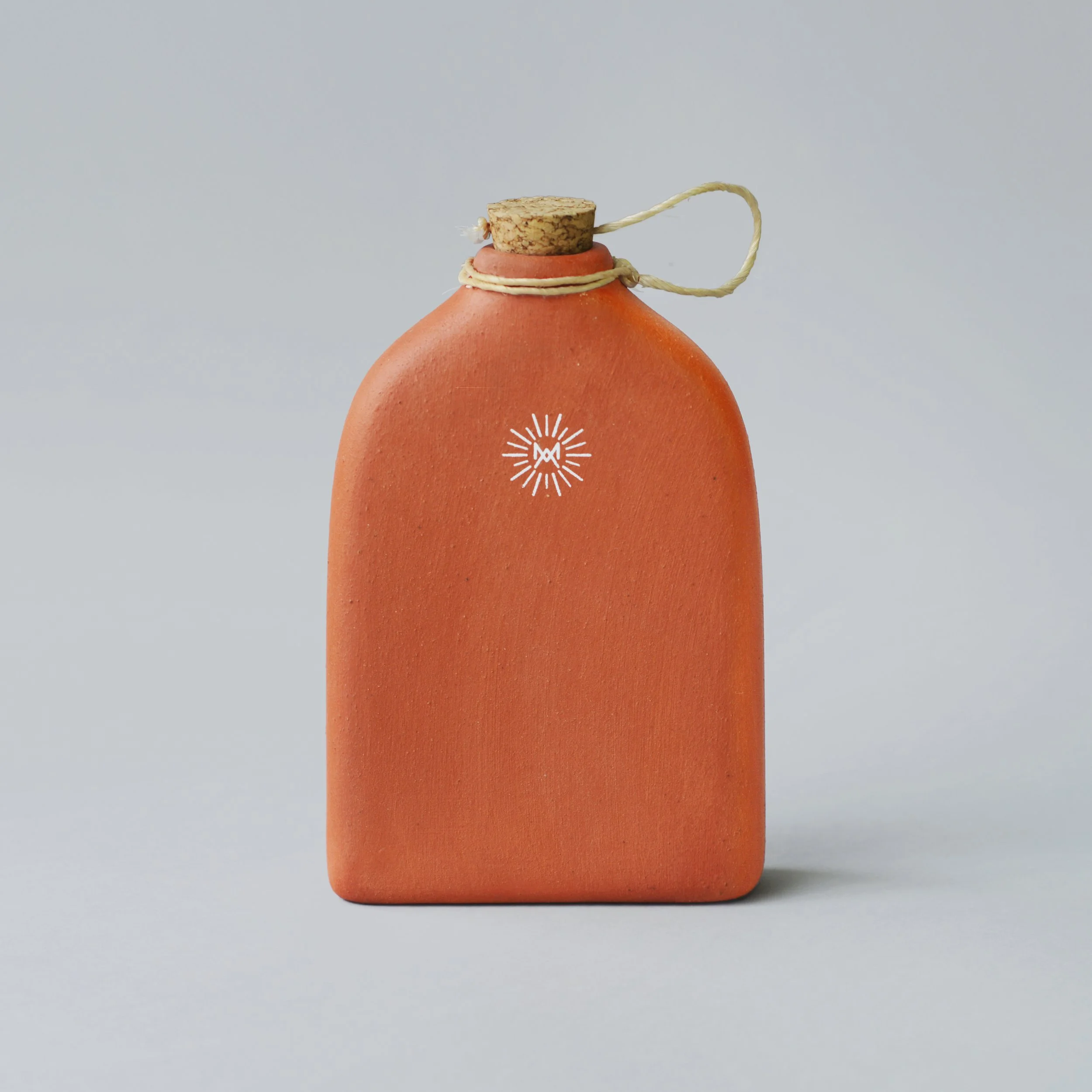 A terracotta-colored ceramic bottle with a cork stopper and a rope loop attached, featuring a white sunburst logo with the letter 'M' at the center.