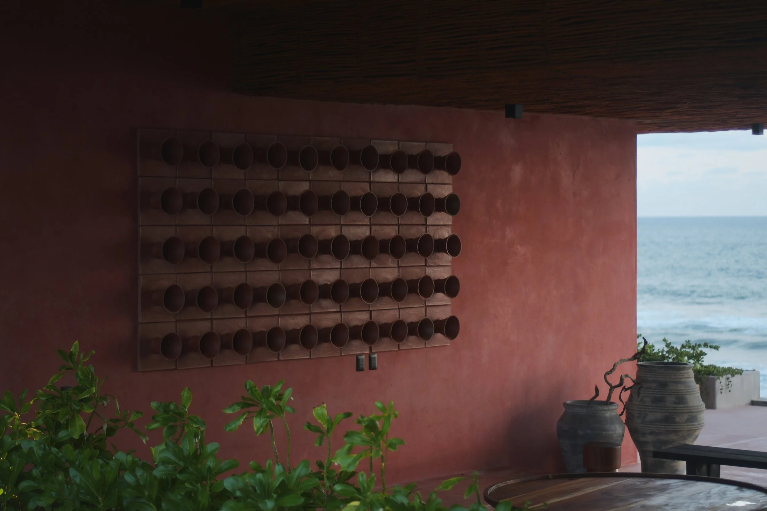 Terracotta pots arranged on a wall with ocean view in background, over a pink wall and wooden roof.