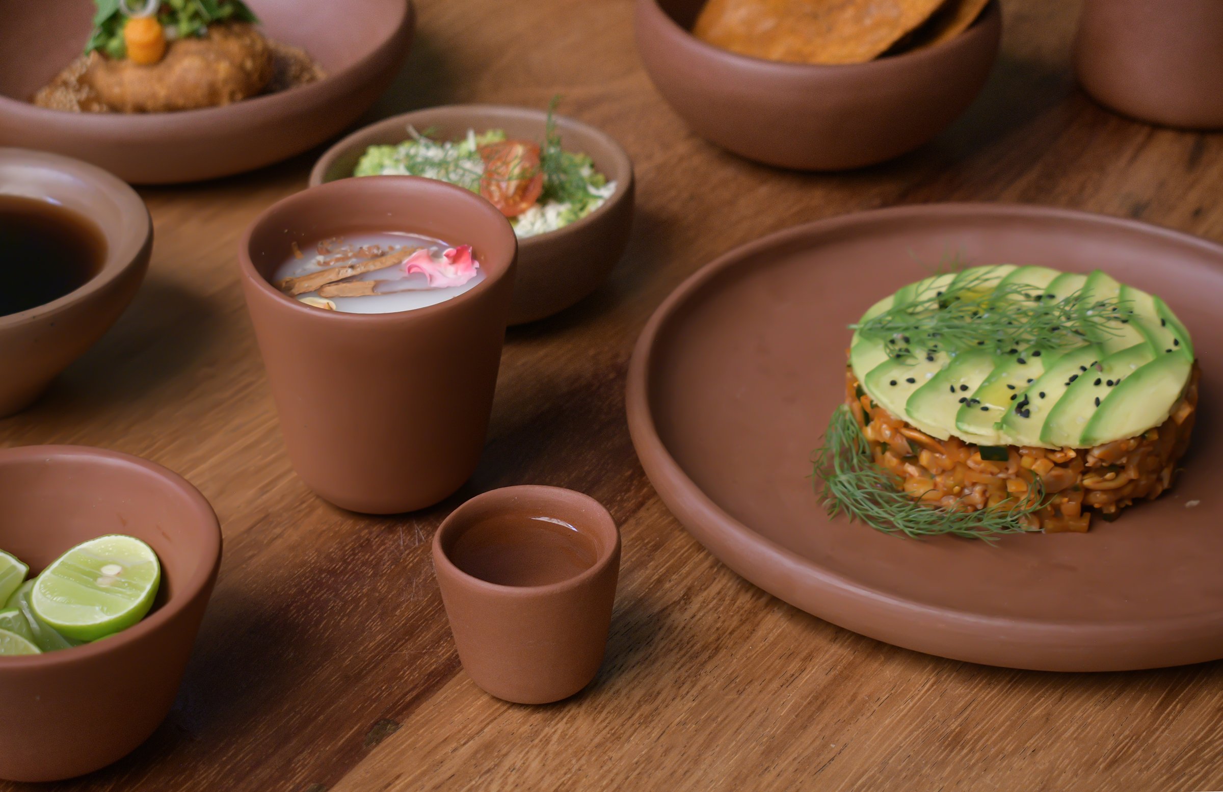 Various bowls of food on a wooden table, including cut lime slices, a dessert with avocado slices and microgreens, a drink with pink flower decoration, a salad, and chips.