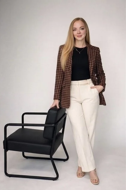 A woman with long blonde hair wearing a plaid blazer, black top, and cream-colored pants, standing next to a modern black chair with armrests against a plain light gray background.