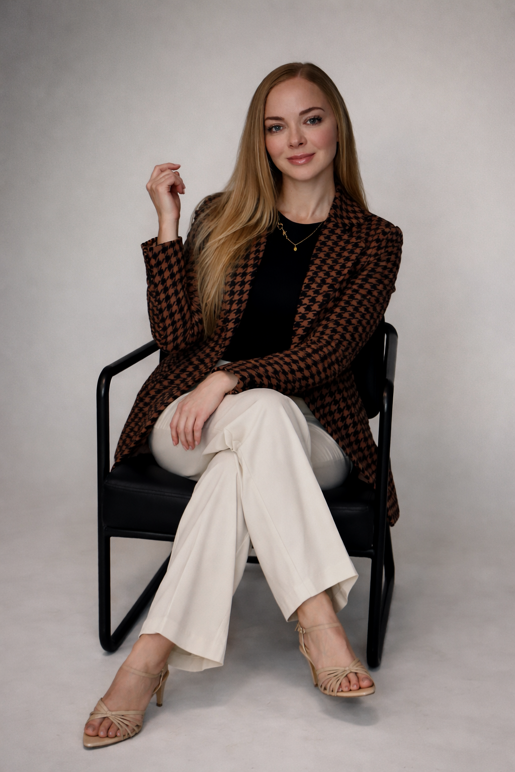 A woman with long blonde hair, wearing a brown houndstooth blazer, black top, white wide-leg pants, and beige high heels, seated on a black chair against a plain light gray background.