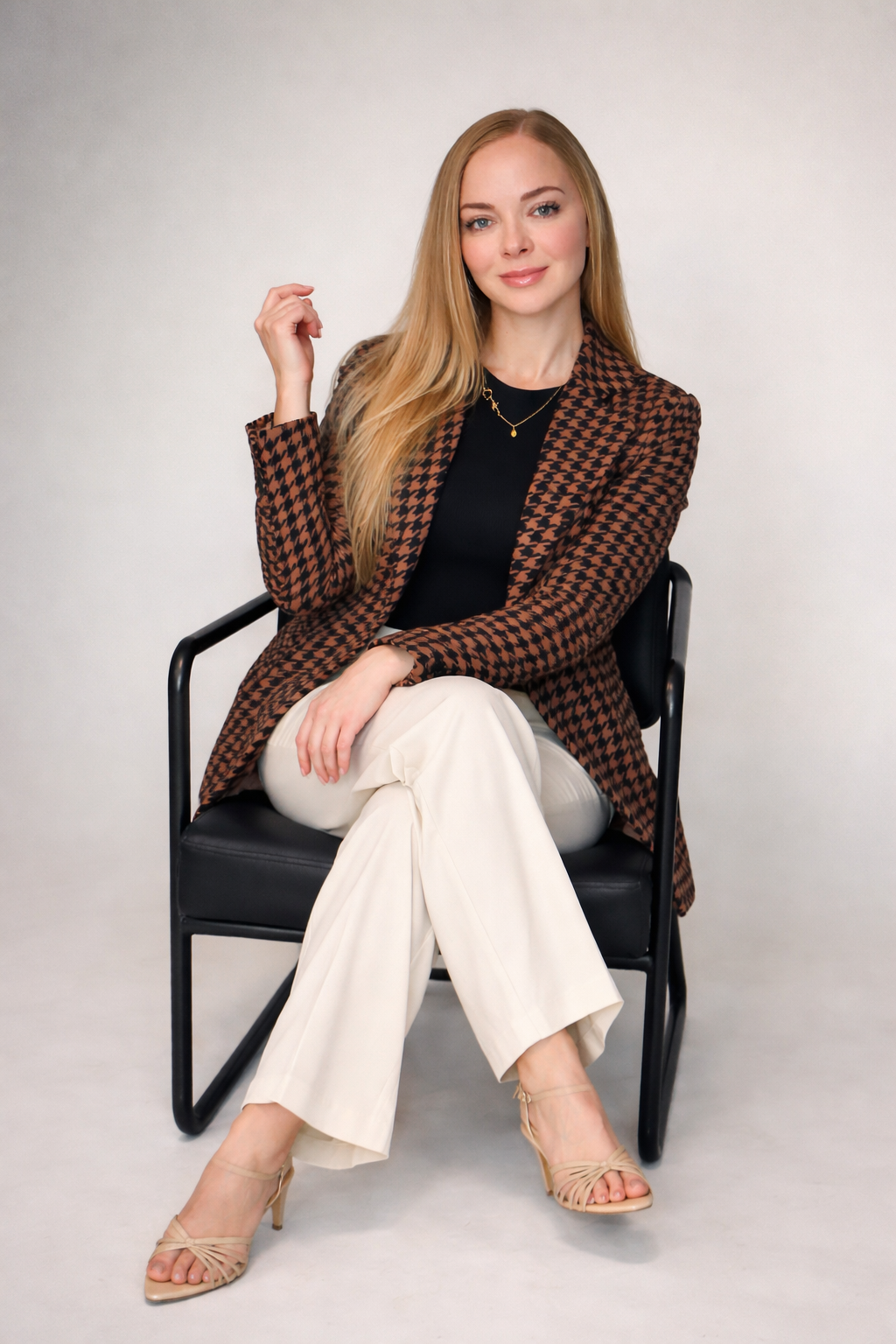 A woman with long blonde hair sitting on a black chair with a light grey background. She is wearing a brown houndstooth blazer, black top, cream-colored pants, and beige high heels. She has a small gold necklace and is smiling slightly.