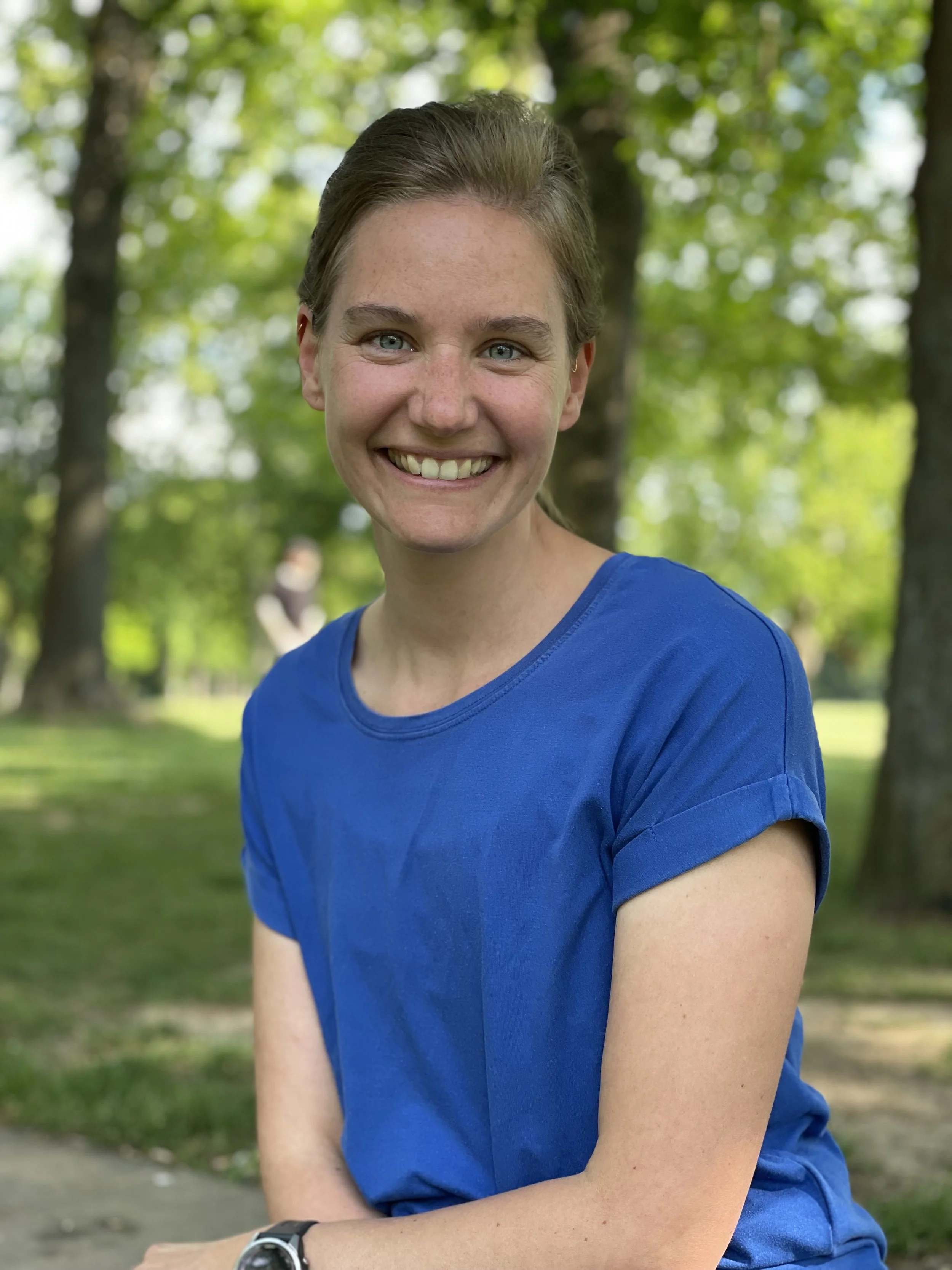 Ein lächelnder junger Mann mit blauen Augen und braunen Haaren, trägt ein blaues T-Shirt und eine Armbanduhr, sitzt im Park mit grünen Bäumen im Hintergrund.
