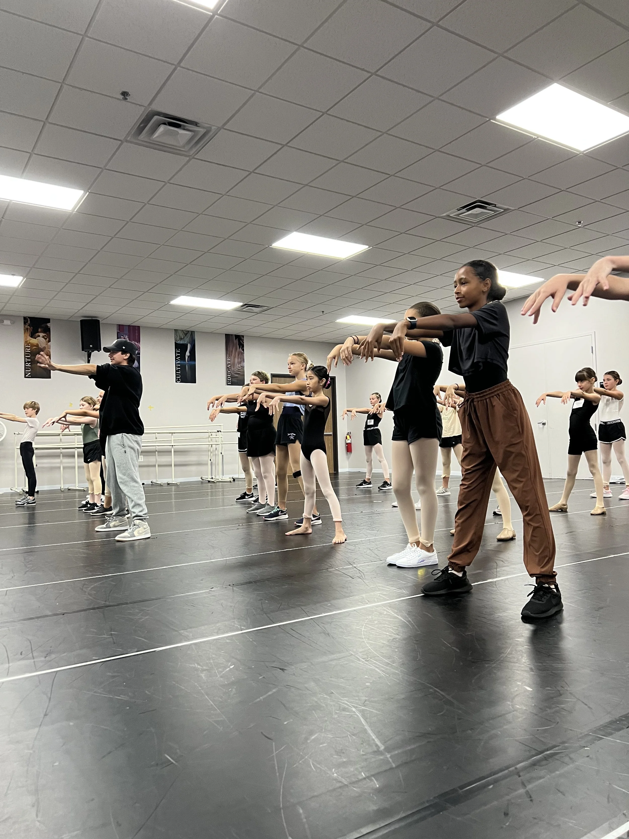Group of young ballet dancers practicing at a dance studio, following dance instructor in black cap and gray sweatpants. Dancers wearing ballet attire, some in leotards and tights, are standing on a black dance floor with ballet bars along the walls.