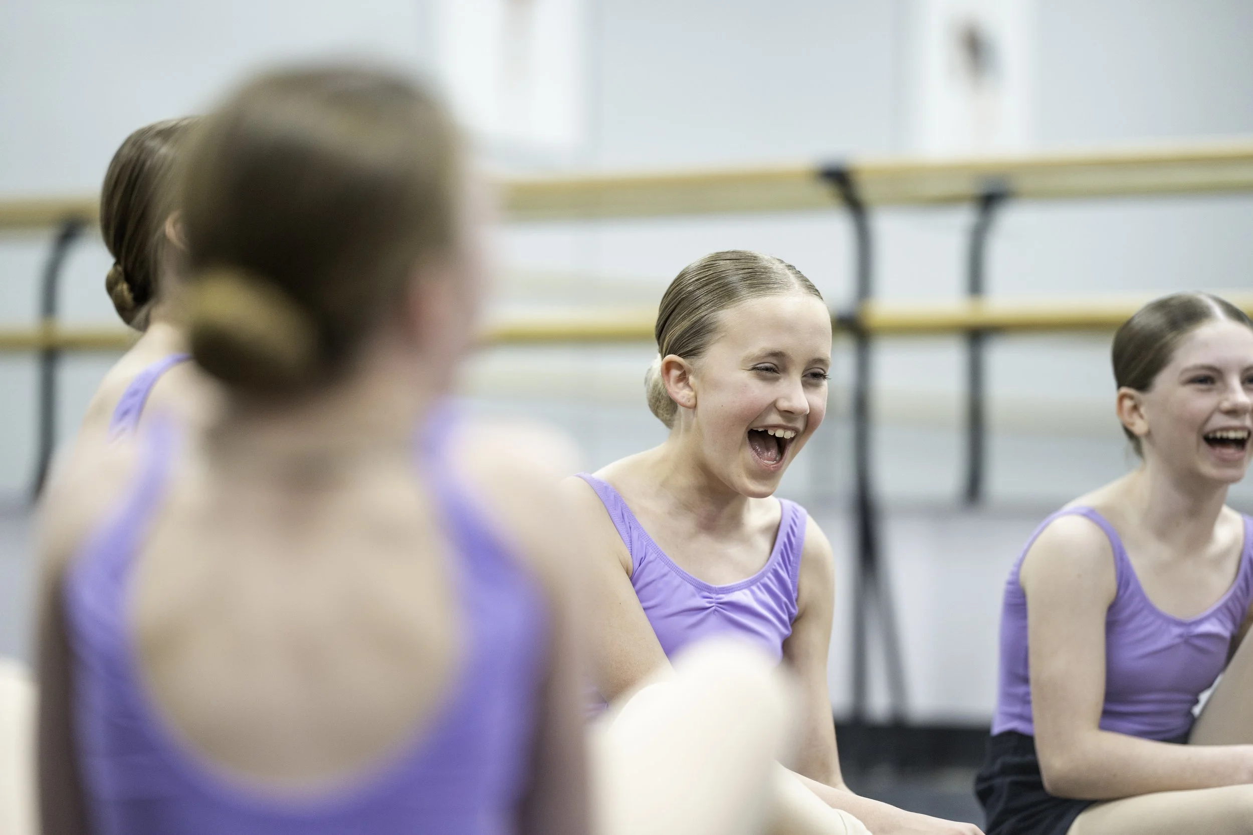 Group of young girls in purple dance costumes sitting and laughing during dance class or rehearsal.