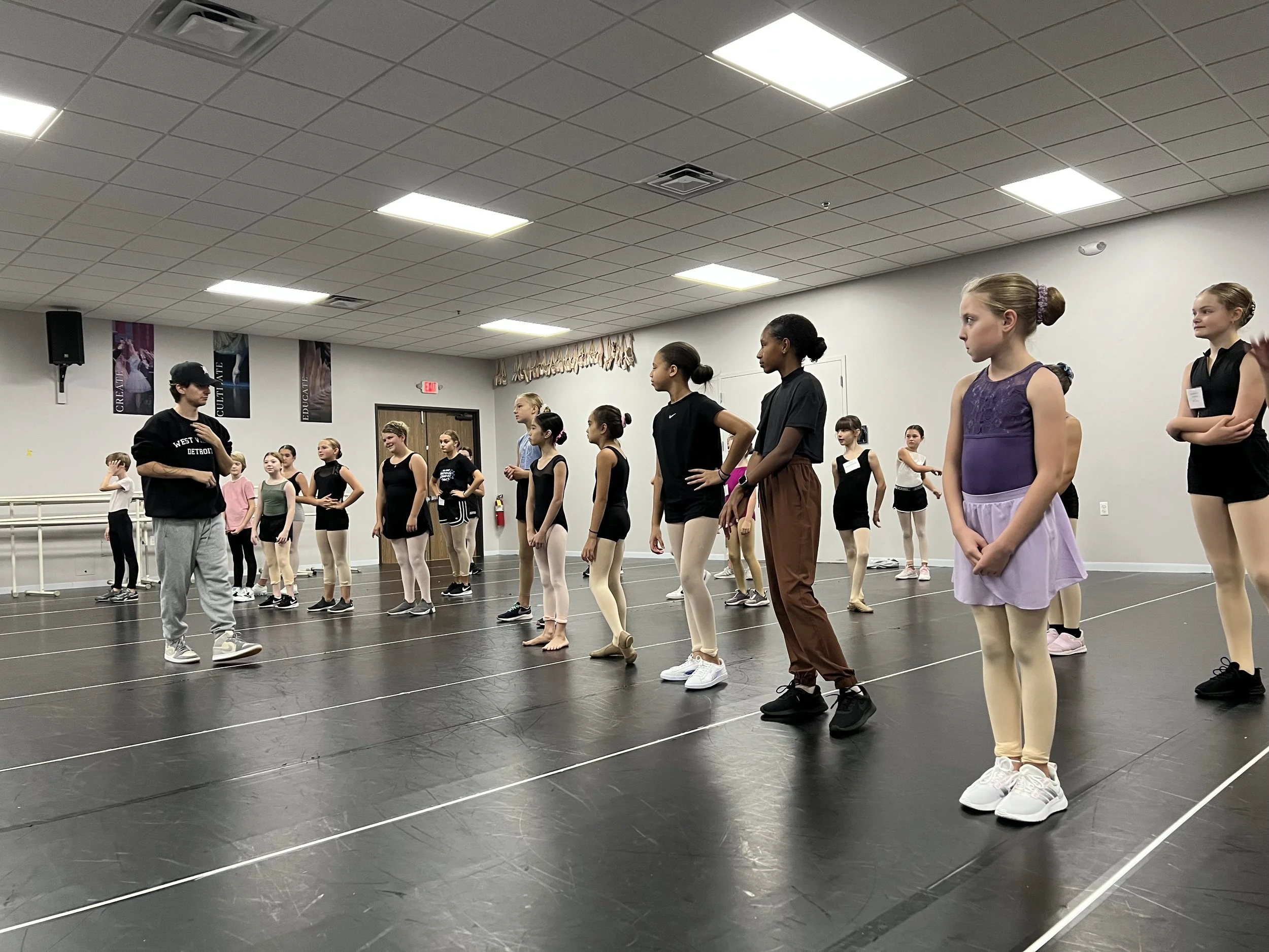 A group of young ballet students standing in a dance studio listening to their instructor, with some wearing leotards and tights, and others in casual dance wear.