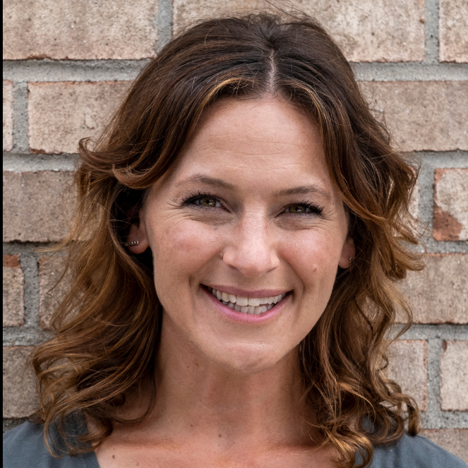 A woman with short brown hair, wearing a gray off-shoulder sweater, smiling and leaning against a white wall.