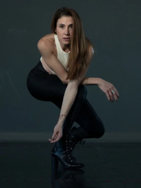 A woman with long hair wearing a sleeveless top and black pants, squatting on the floor, adjusting her shoe.