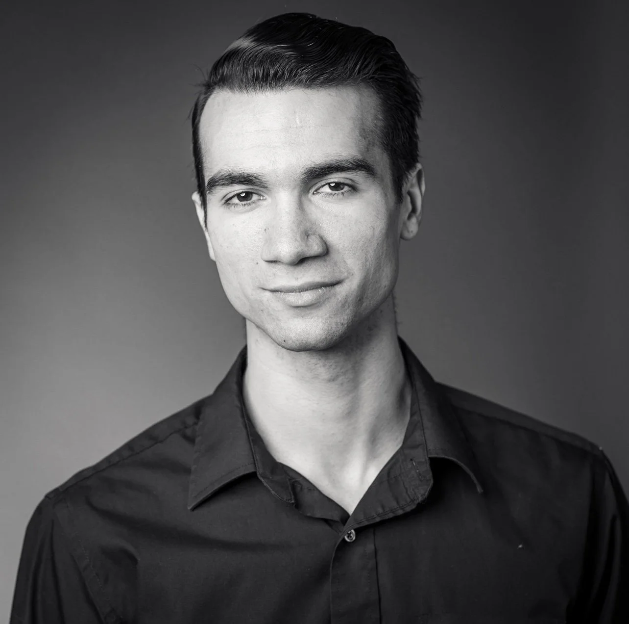 Black and white portrait of a young man with slicked-back hair wearing a dark collared shirt, smiling subtly.