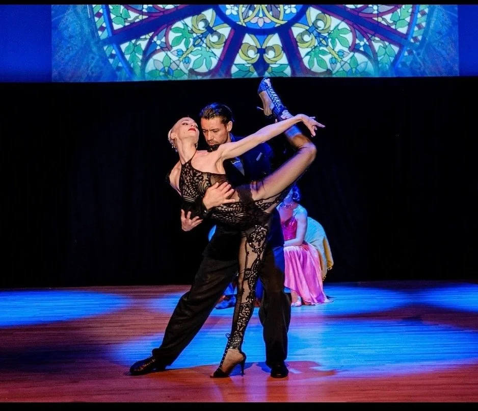 A man and woman dance tango on stage, with the woman lifted in a pose; the woman is wearing a black lace dress and high heels, the man is in black vest and trousers, with a stained-glass window projected above them.