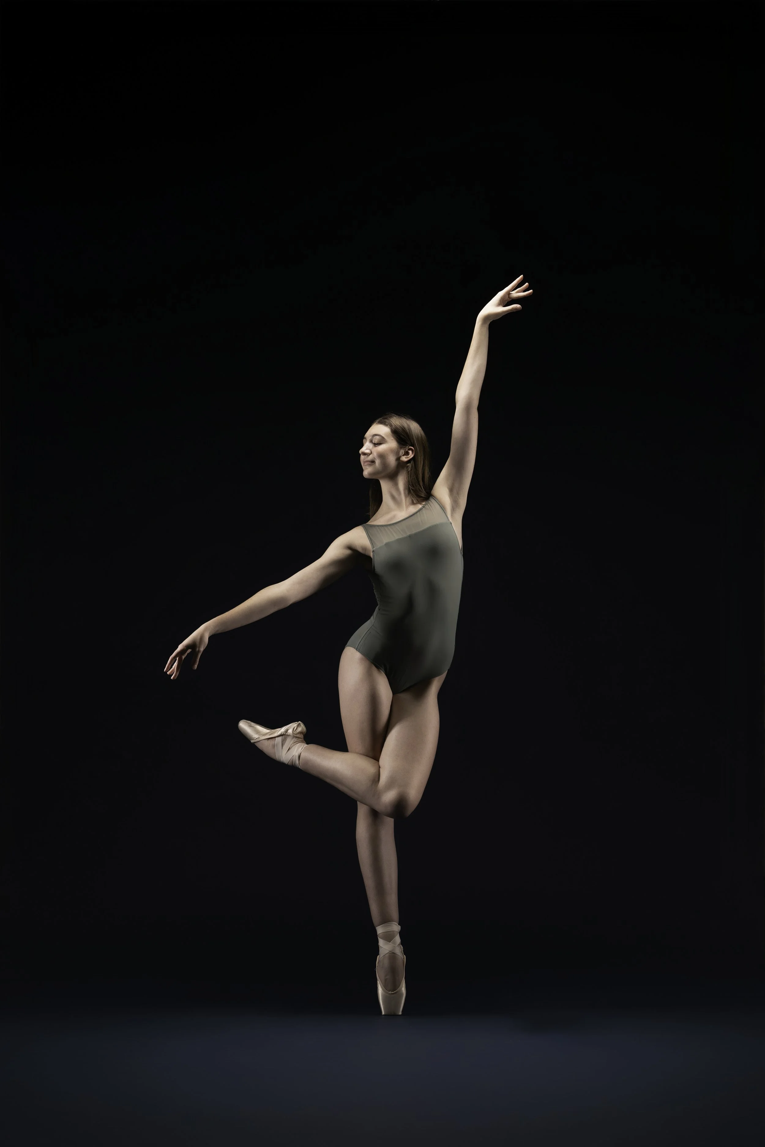 A ballerina performing a dance pose in front of a black background, wearing a green leotard and ballet slippers.