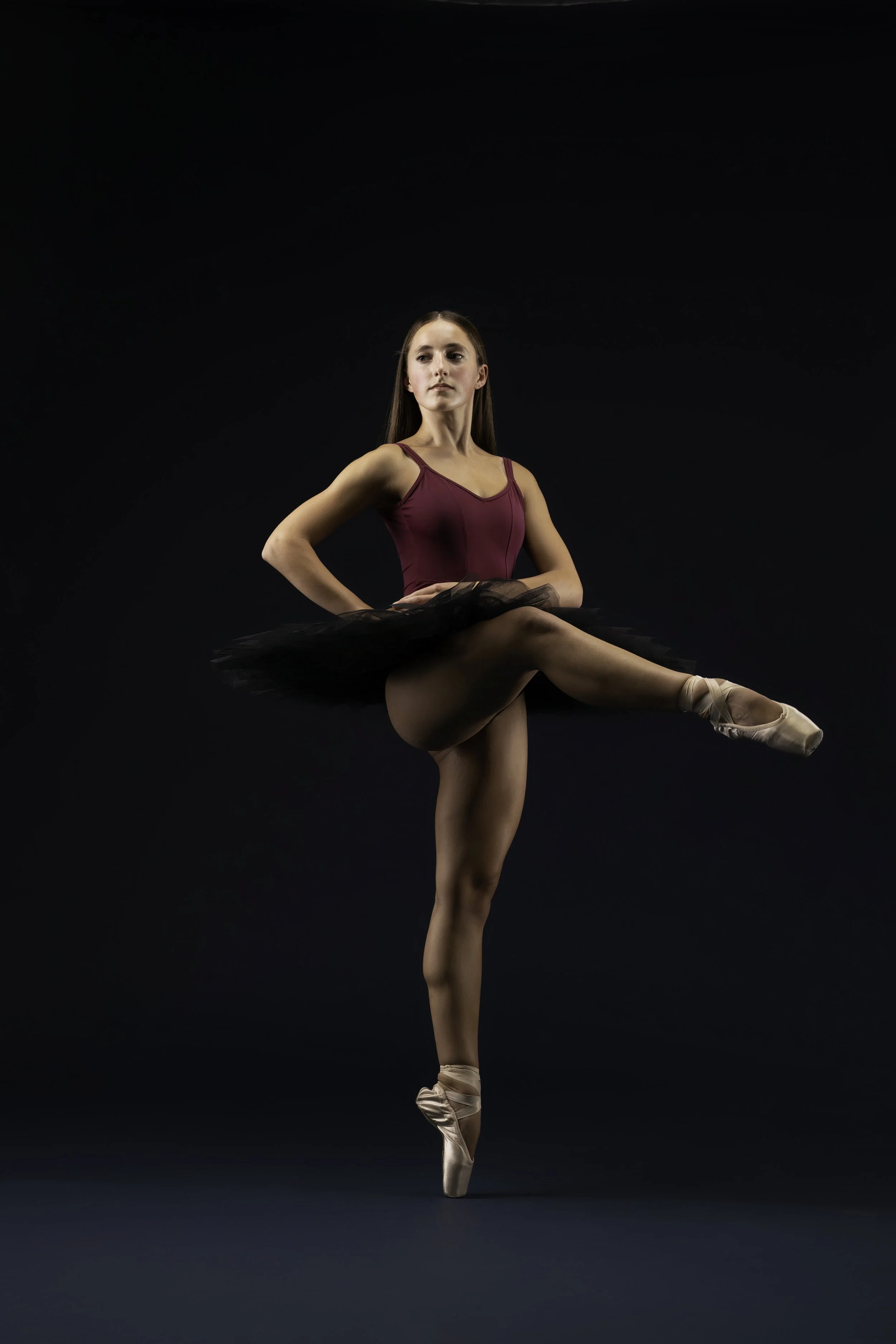A ballet dancer in a burgundy leotard and black tutu, standing on one leg with the other leg extended and pointed, wearing pink ballet slippers, against a dark background.