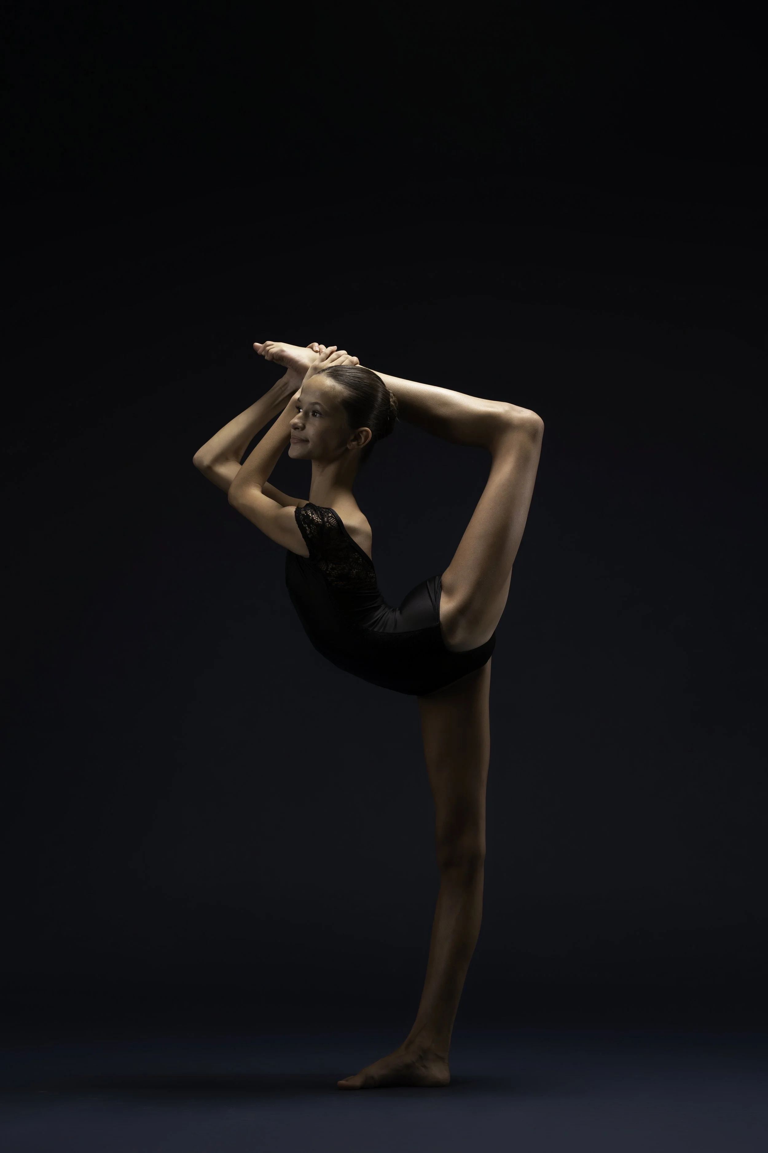 A young woman performing a yoga pose called Natarajasana or Lord of the Dance Pose, on a dark background.