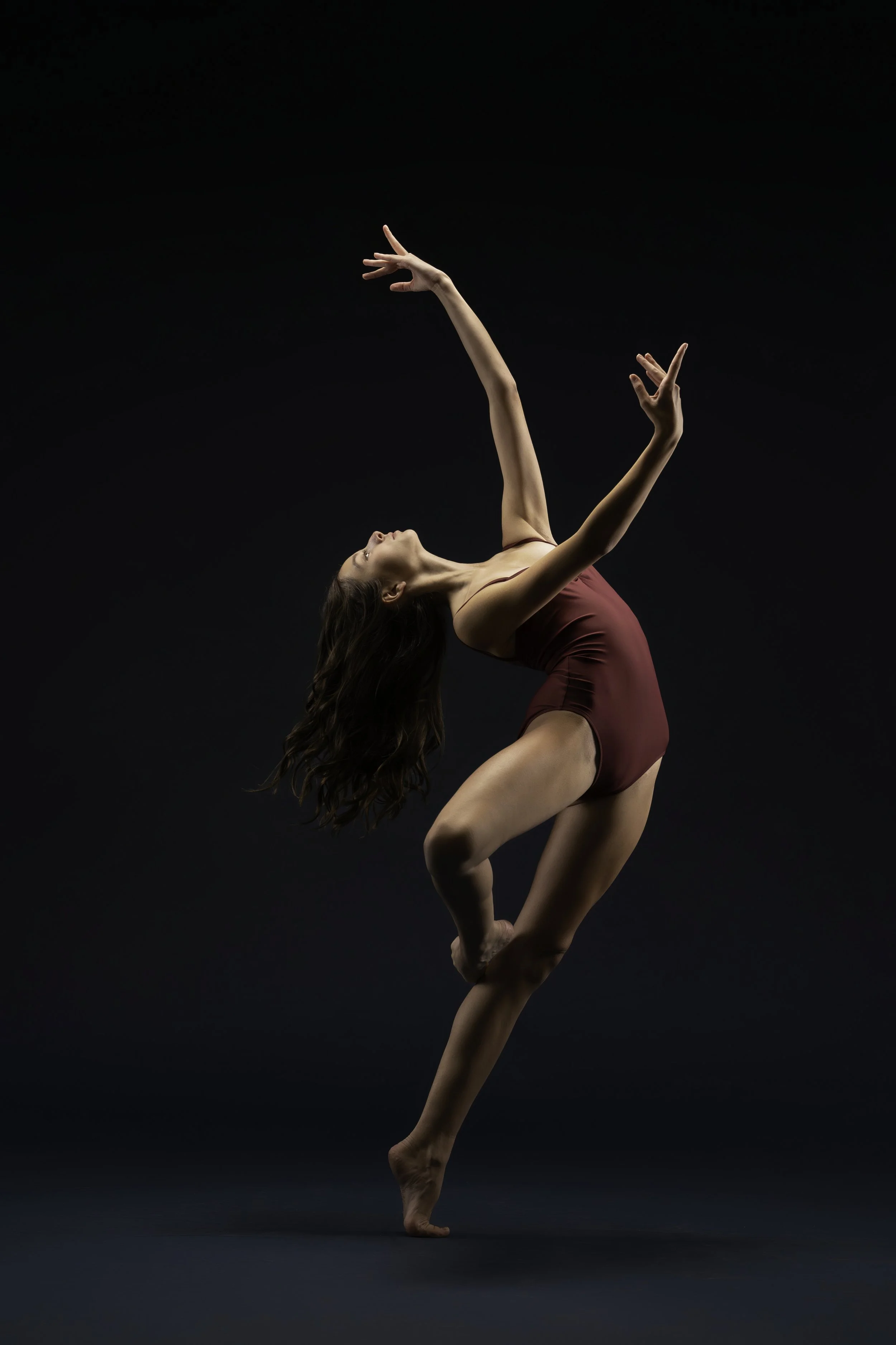 A female dancer performing a contemporary dance move against a black background, wearing a maroon leotard, with her body arched and arms extended gracefully.