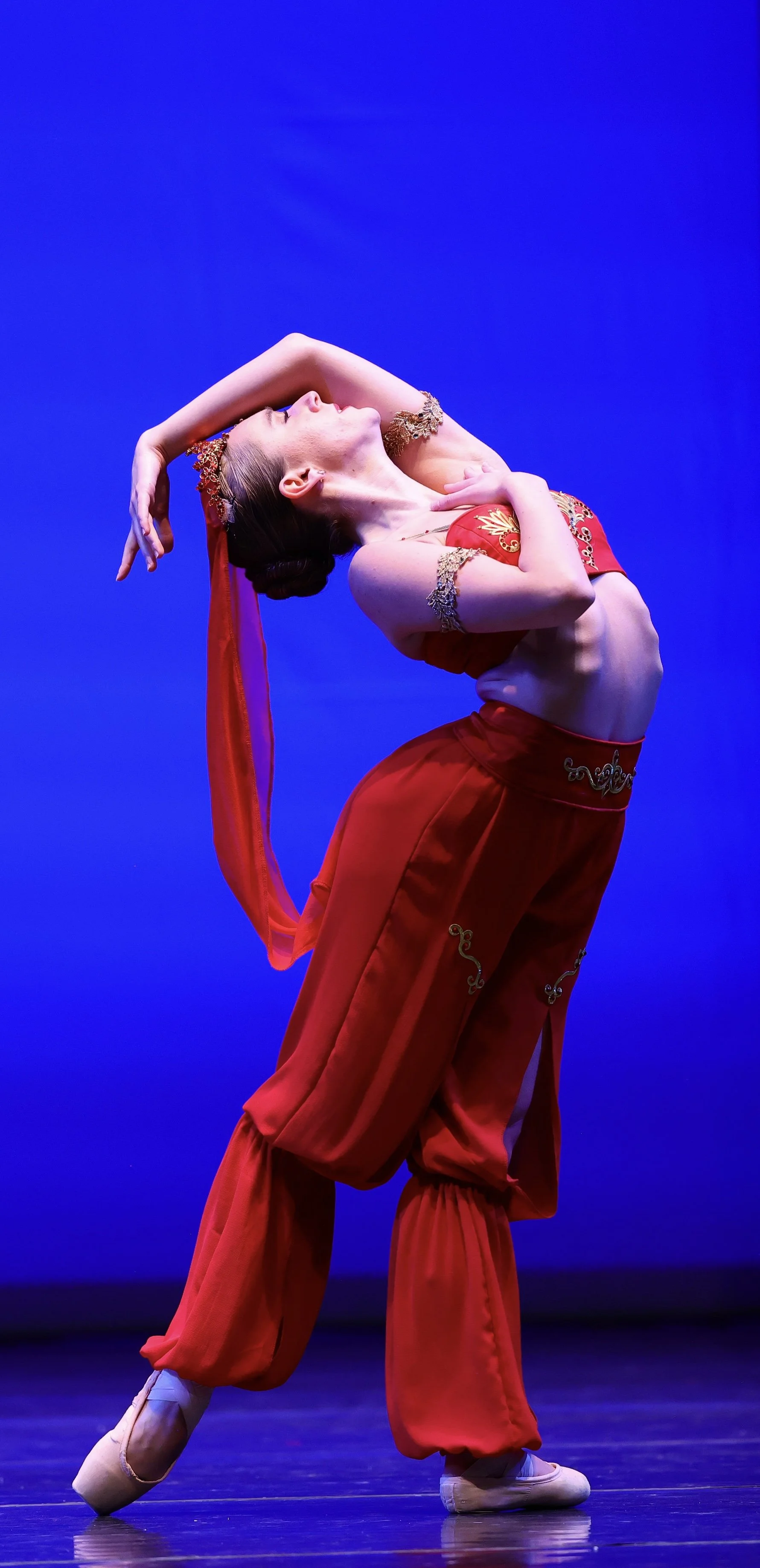 A ballet dancer in a red costume performing a pose on stage with a blue background.