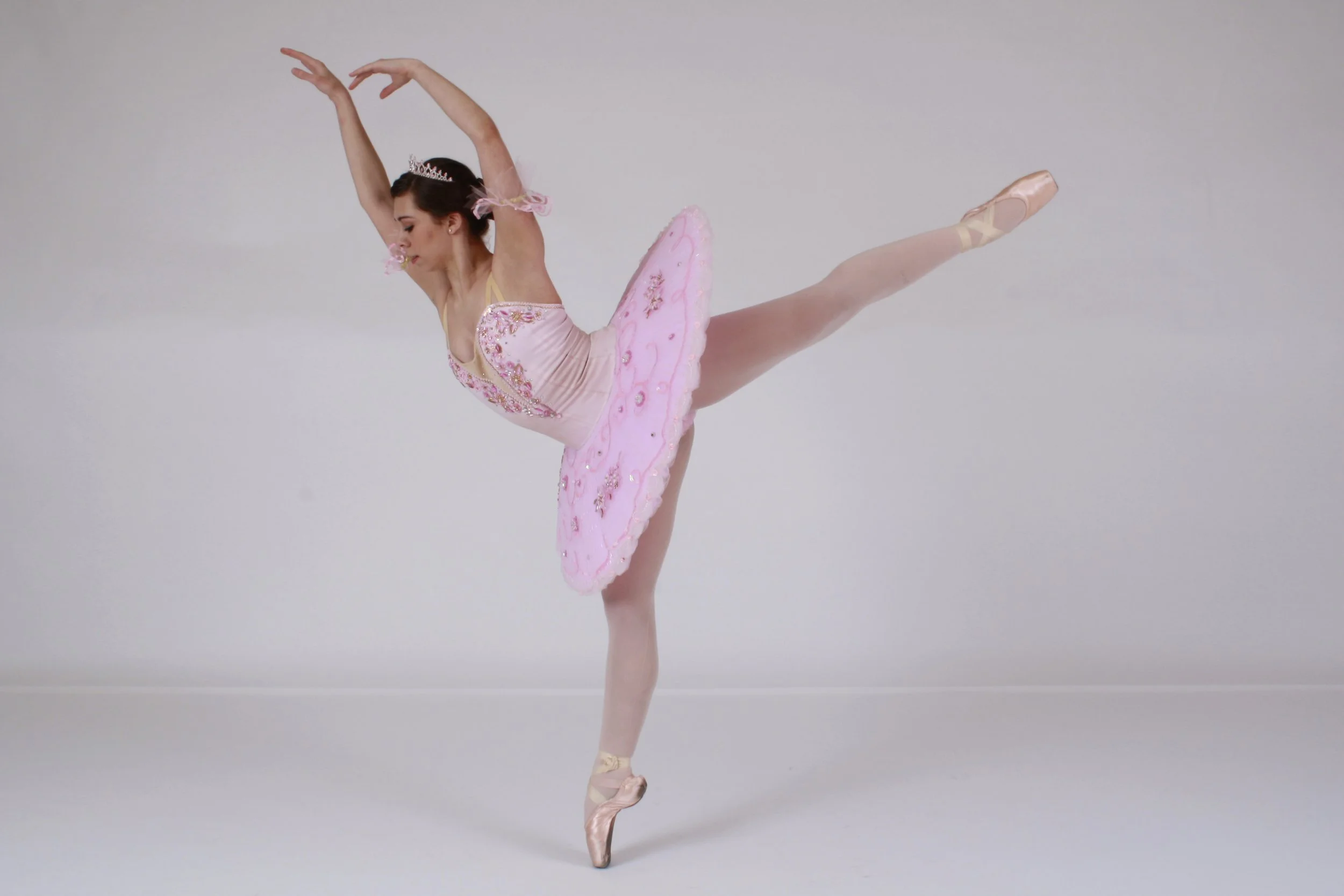 Ballet dancer in pink tutu performing an arabesque pose, standing on one foot with arms curved overhead.