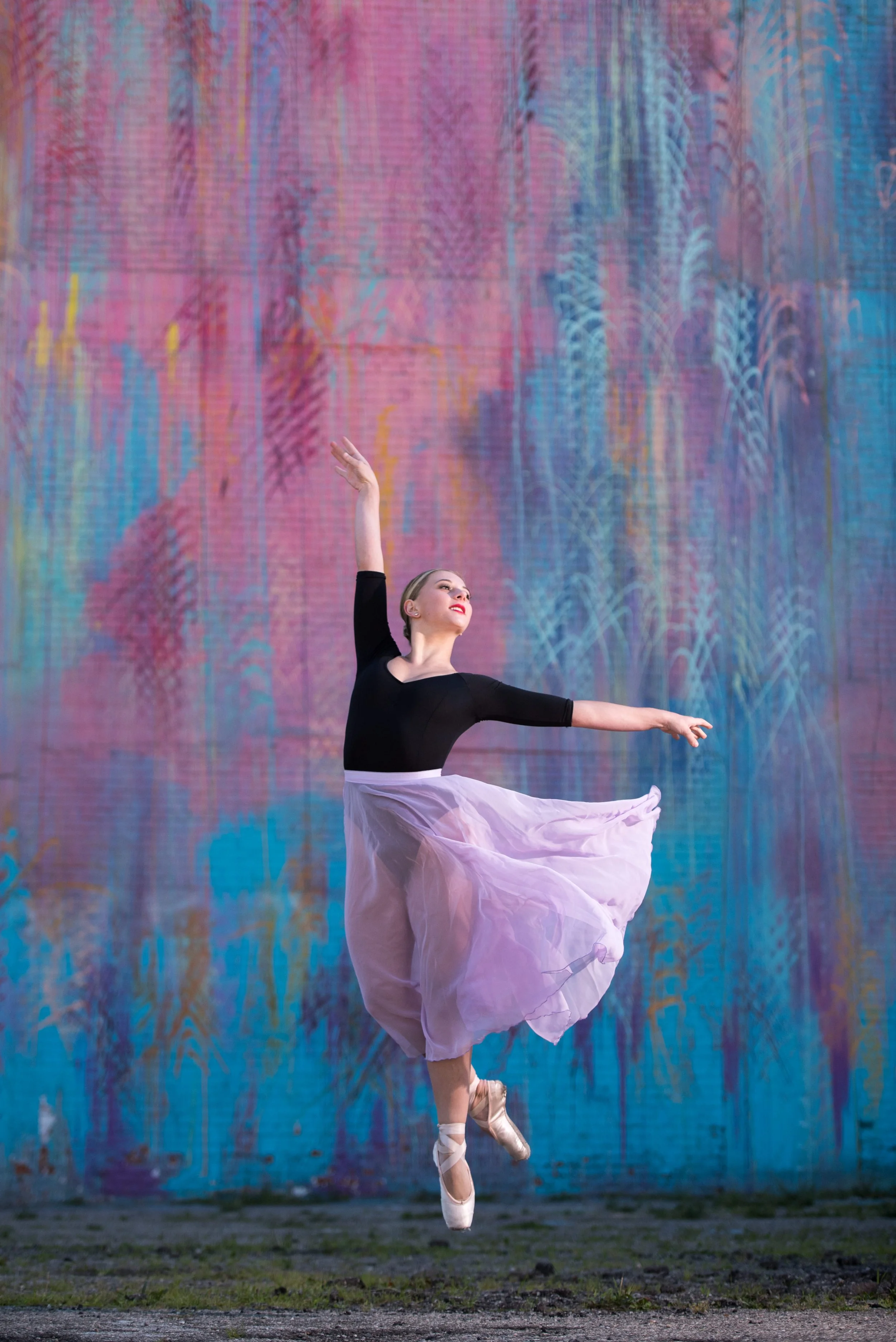 A female ballet dancer in a black top and a flowing pink skirt performing a leap on pointe in front of a colorful, abstract mural wall.
