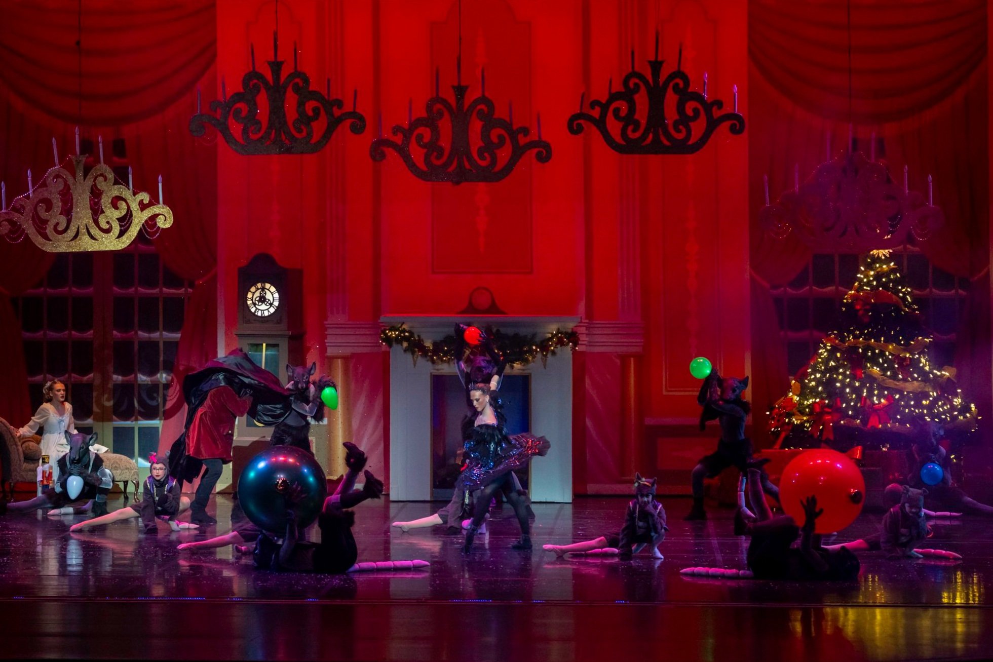 Children performing a holiday ballet on stage decorated for Christmas, with a Christmas tree, garland, and large colorful balls.