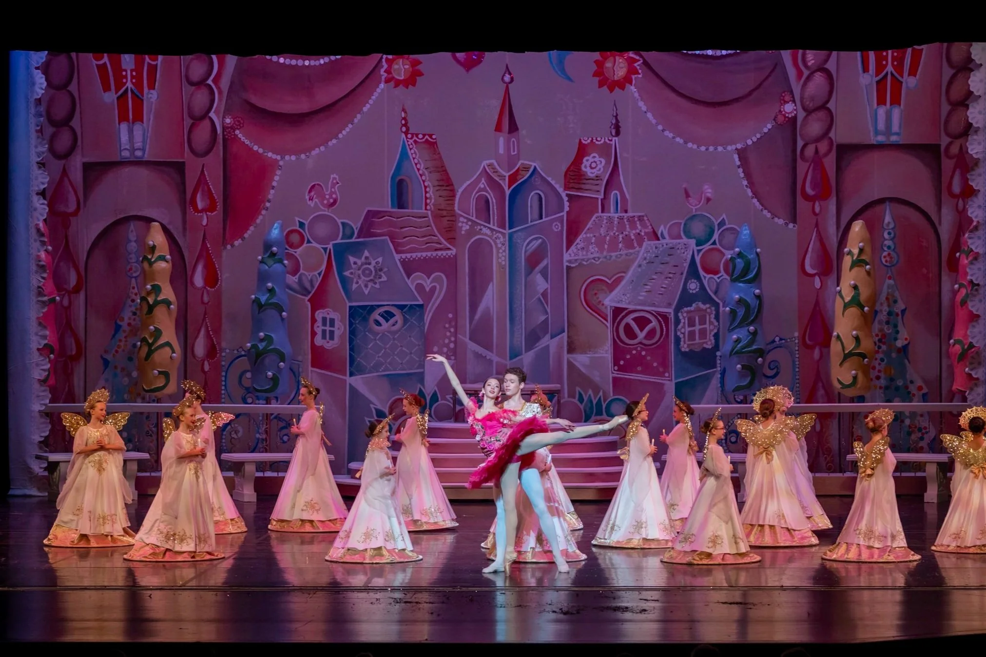 Ballet dancers performing on stage with a colorful whimsical backdrop of a fairy tale village and decorated trees, all dressed in pastel costumes with some wearing fairy wings.