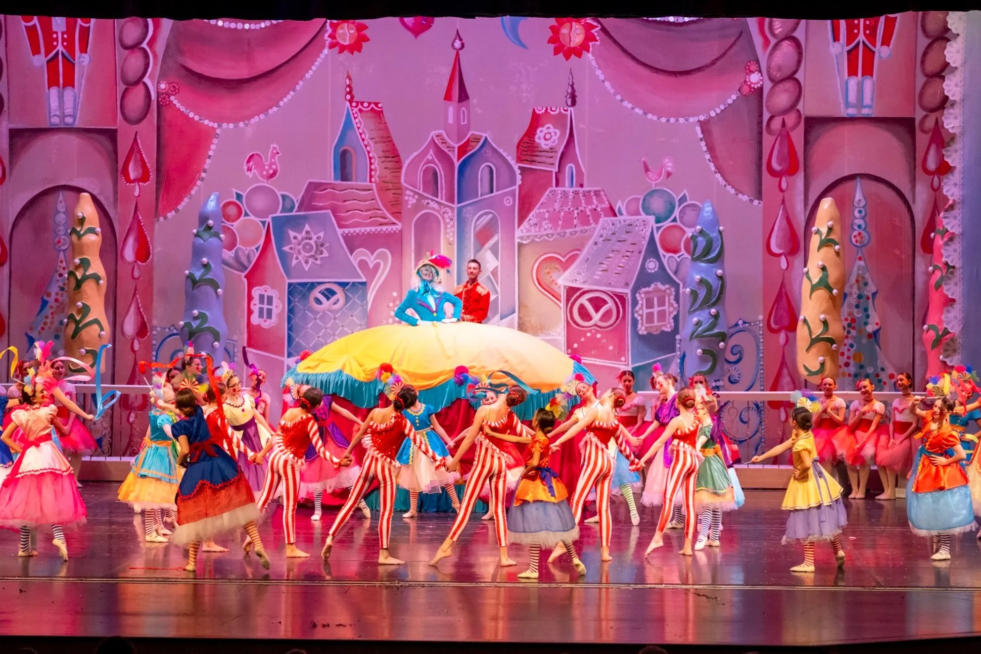 A stage performance with dancers in colorful costumes, a large yellow skirted female character on a raised platform, and a stylized pink and purple castle backdrop with whimsical houses and trees.