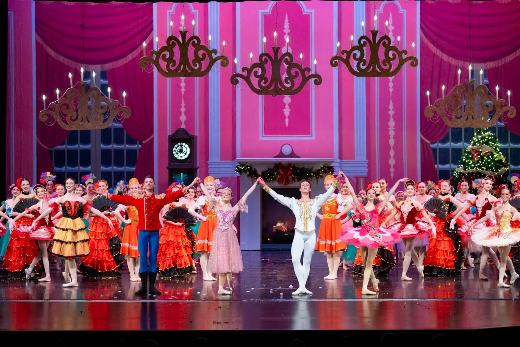 Ballet dancers performing on stage with pink and red Christmas-themed decorations, including a decorated Christmas tree and festive garland, during a holiday ballet show.