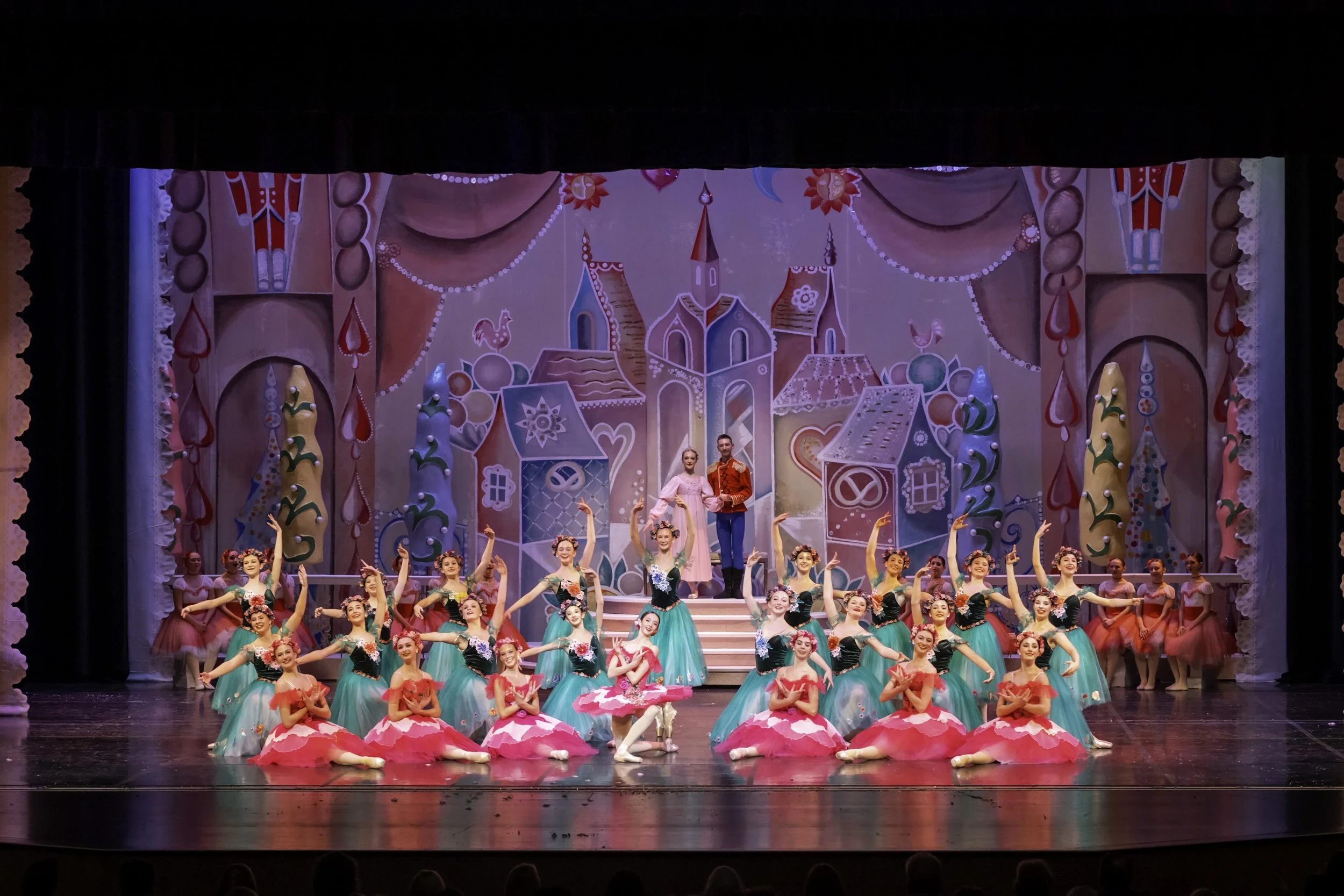 Ballet dancers in colorful costumes performing on stage with a fairy tale backdrop of houses, trees, and a castle in pinks and greens.