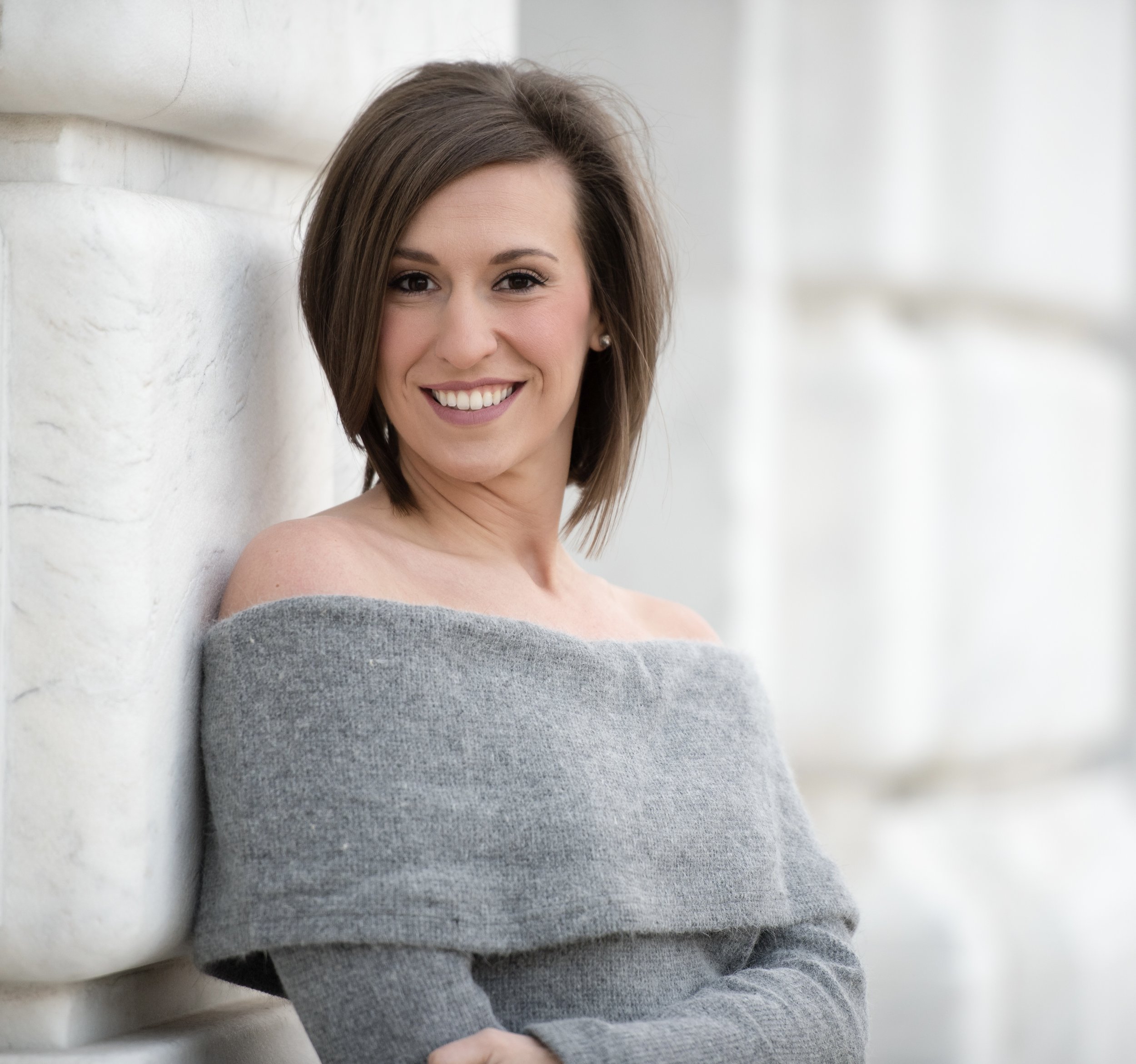 A smiling woman with shoulder-length brown hair leaning against a white stone wall, wearing an off-the-shoulder gray sweater.