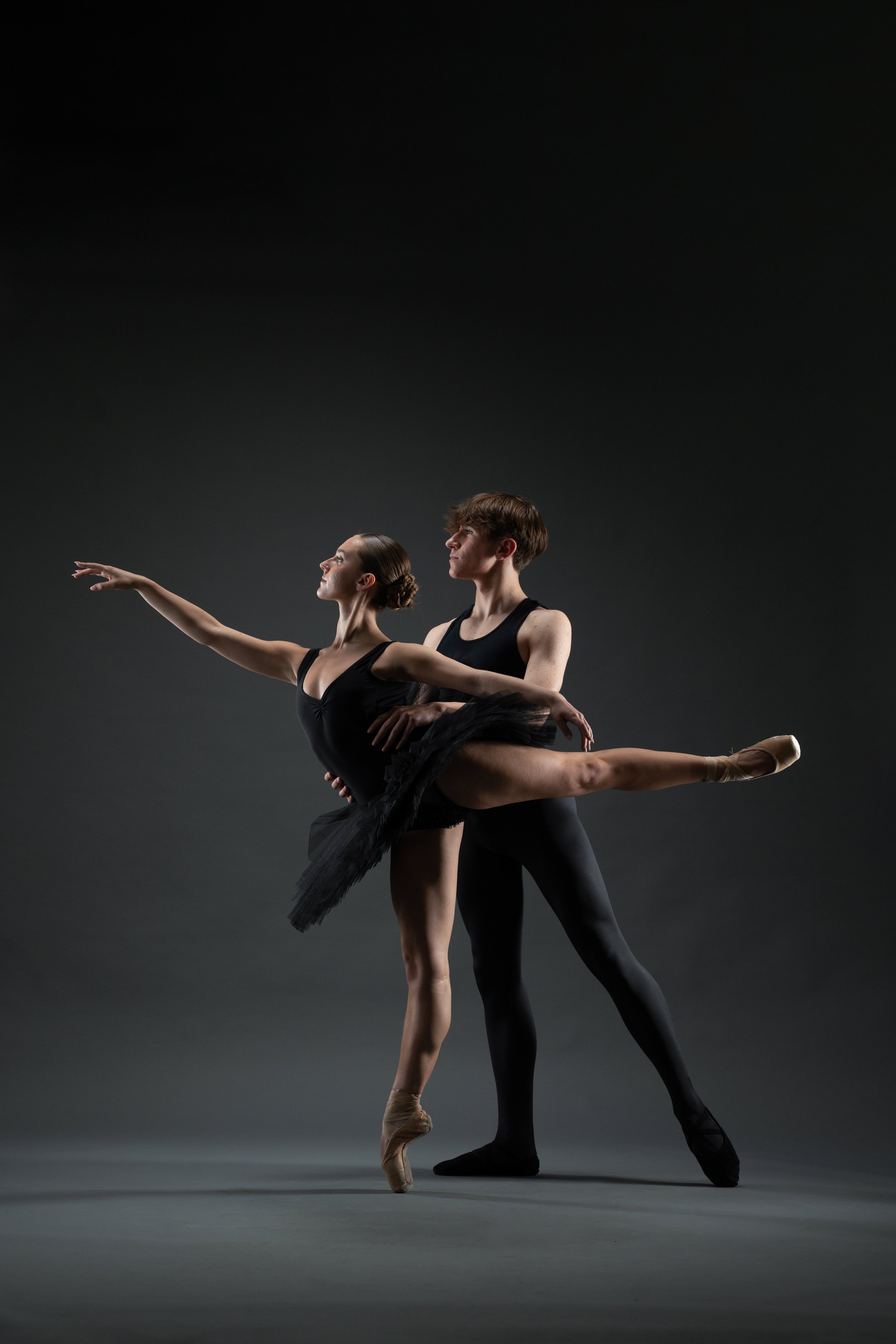 A ballerina and a male dancer performing a dance pose in a dark studio, with the female dancer on pointe in a black tutu, extended leg, and arms, supported by the male dancer.