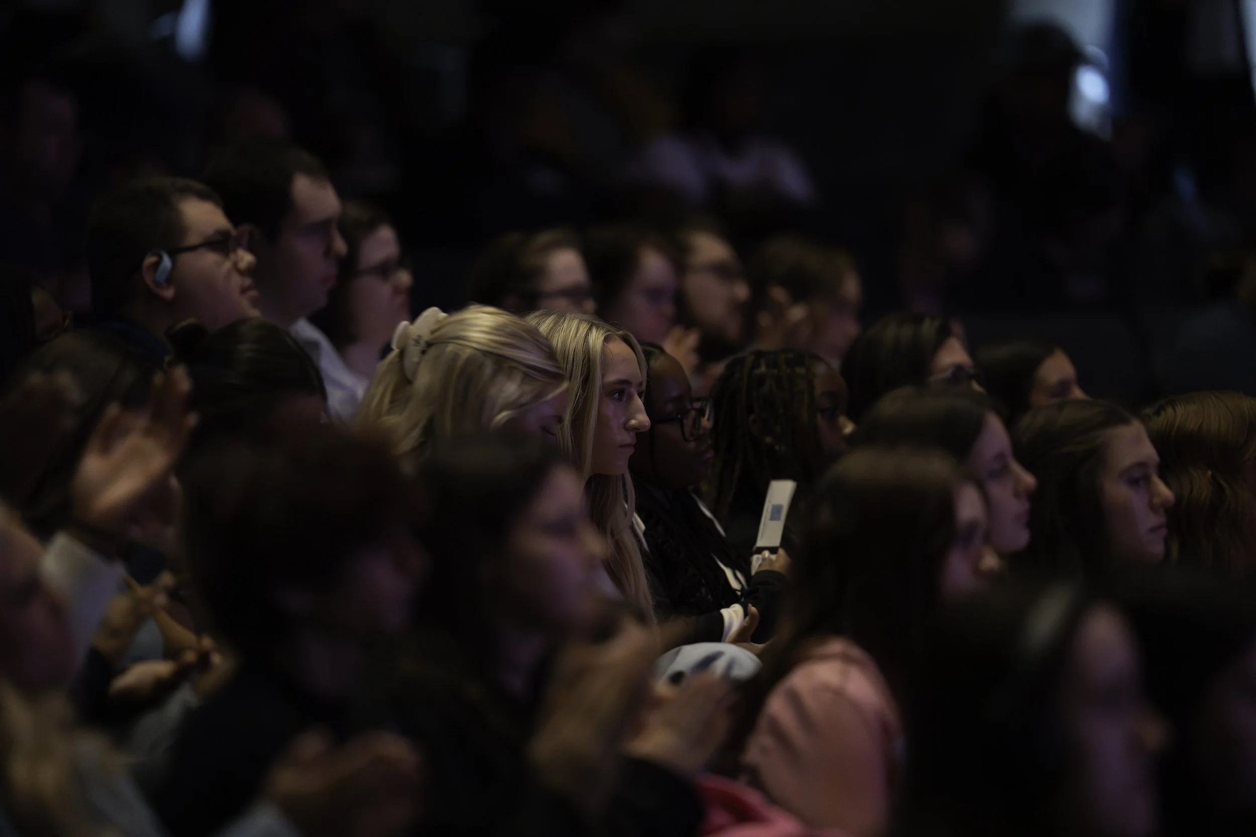 Audience of young adults listening attentively at an event in a dark auditorium.