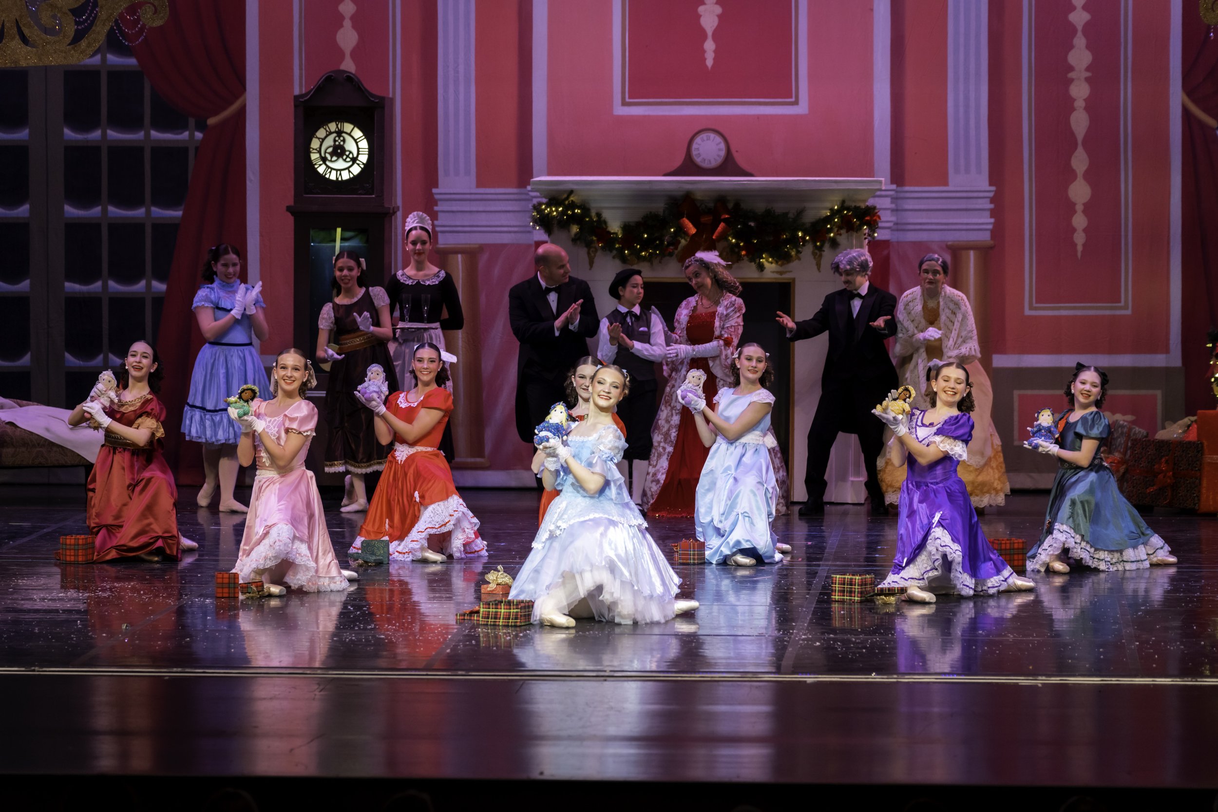 Stage performance of children in colorful Christmas costumes, with adults in formal attire, in a decorated winter holiday scene.