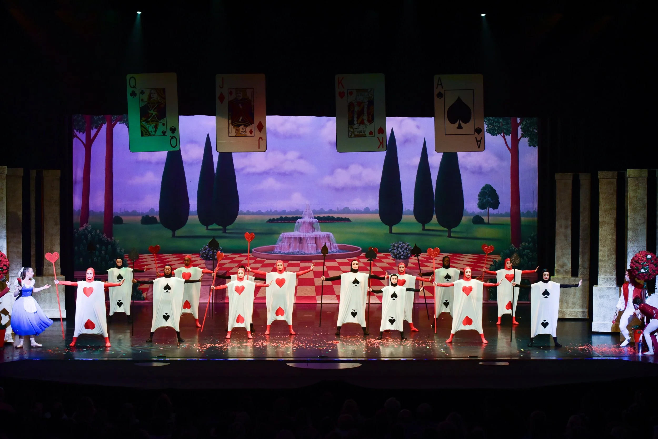 Theatrical stage performance of 'Alice in Wonderland' with dancers dressed as playing cards, set against a colorful backdrop featuring trees, a fountain, and playing cards overhead.