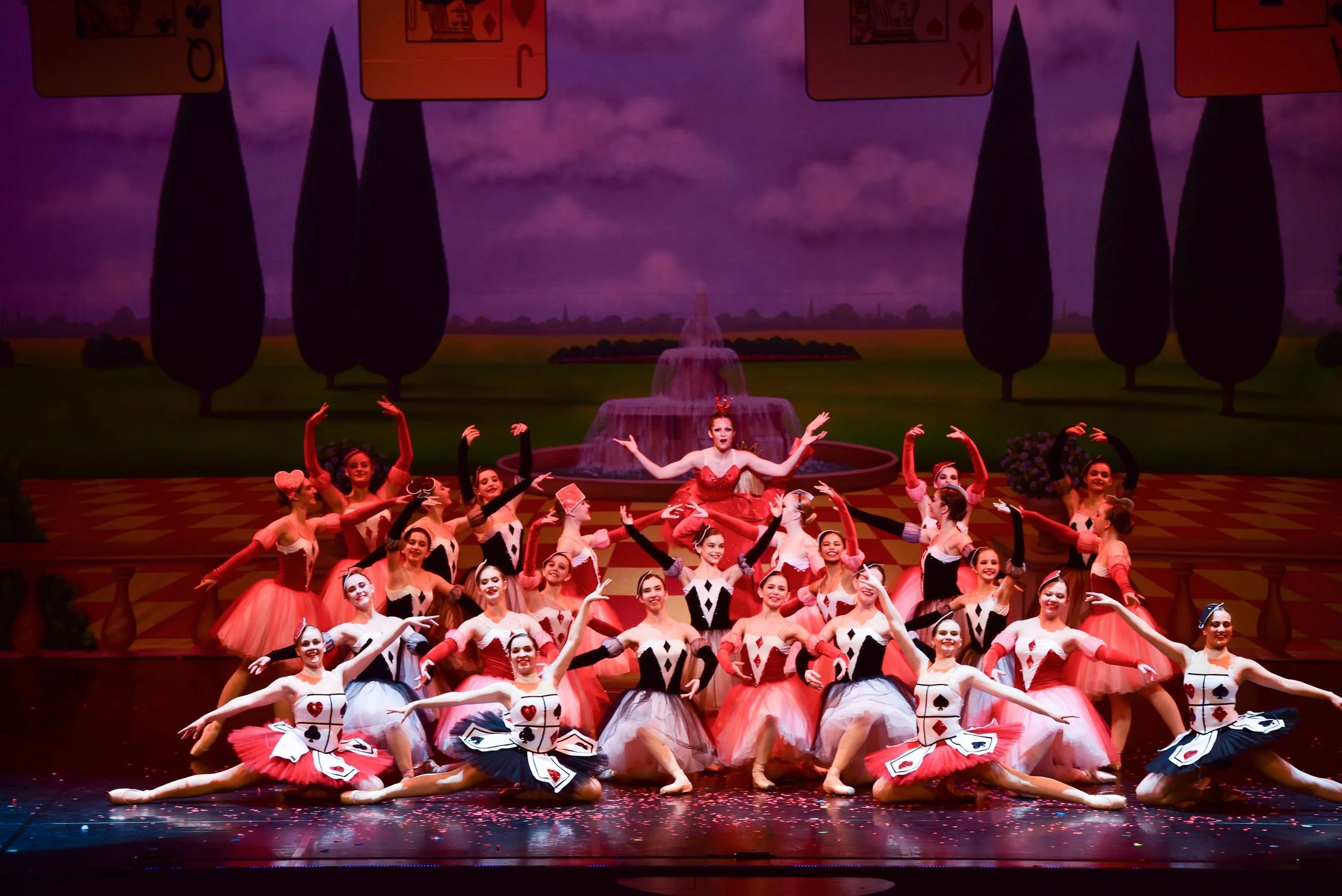 Ballet dancers performing on stage dressed as playing cards, with a backdrop of scenic countryside and a fountain.
