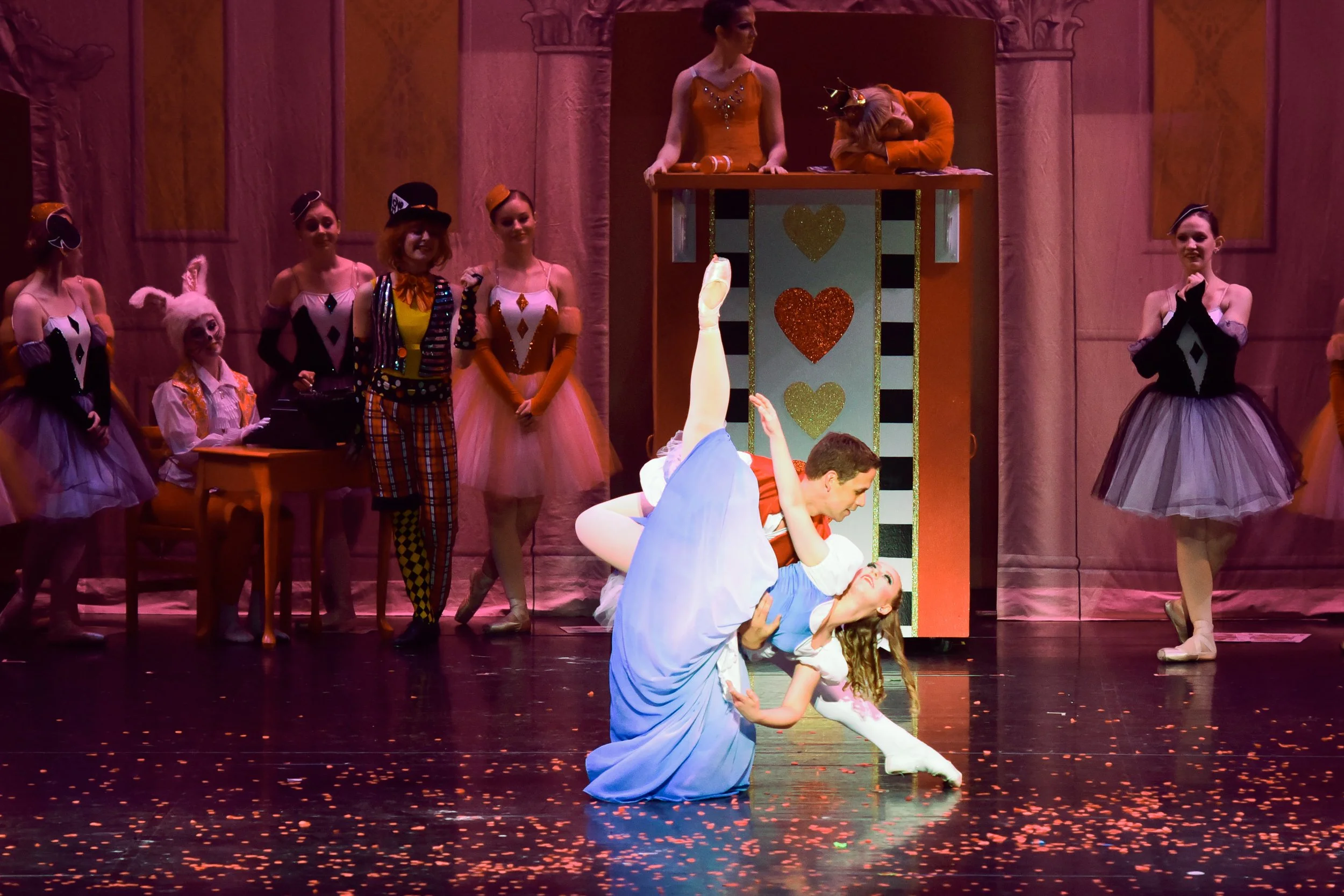 Two ballet dancers performing on stage, with one holding the other in a dip. The stage is decorated with a whimsical, colorful set featuring a tall structure with hearts and stripes, and several performers dressed in colorful costumes and tutus watch