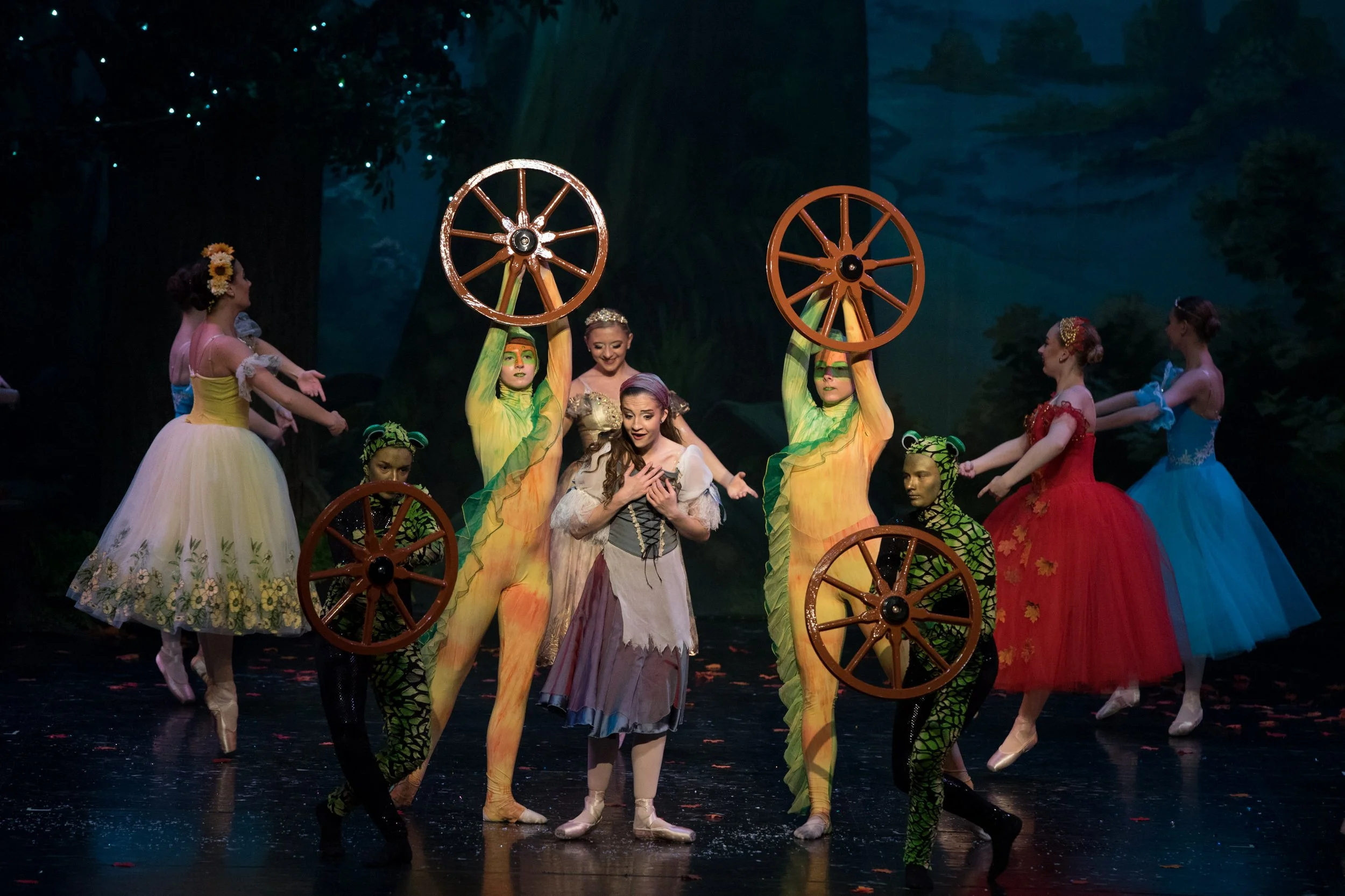 A ballet performance featuring dancers in colorful costumes, including women in flower and sky-colored dresses, and others dressed as animals with green and black patterns, with some holding large wooden wheels, on a stage with a painted forest backg