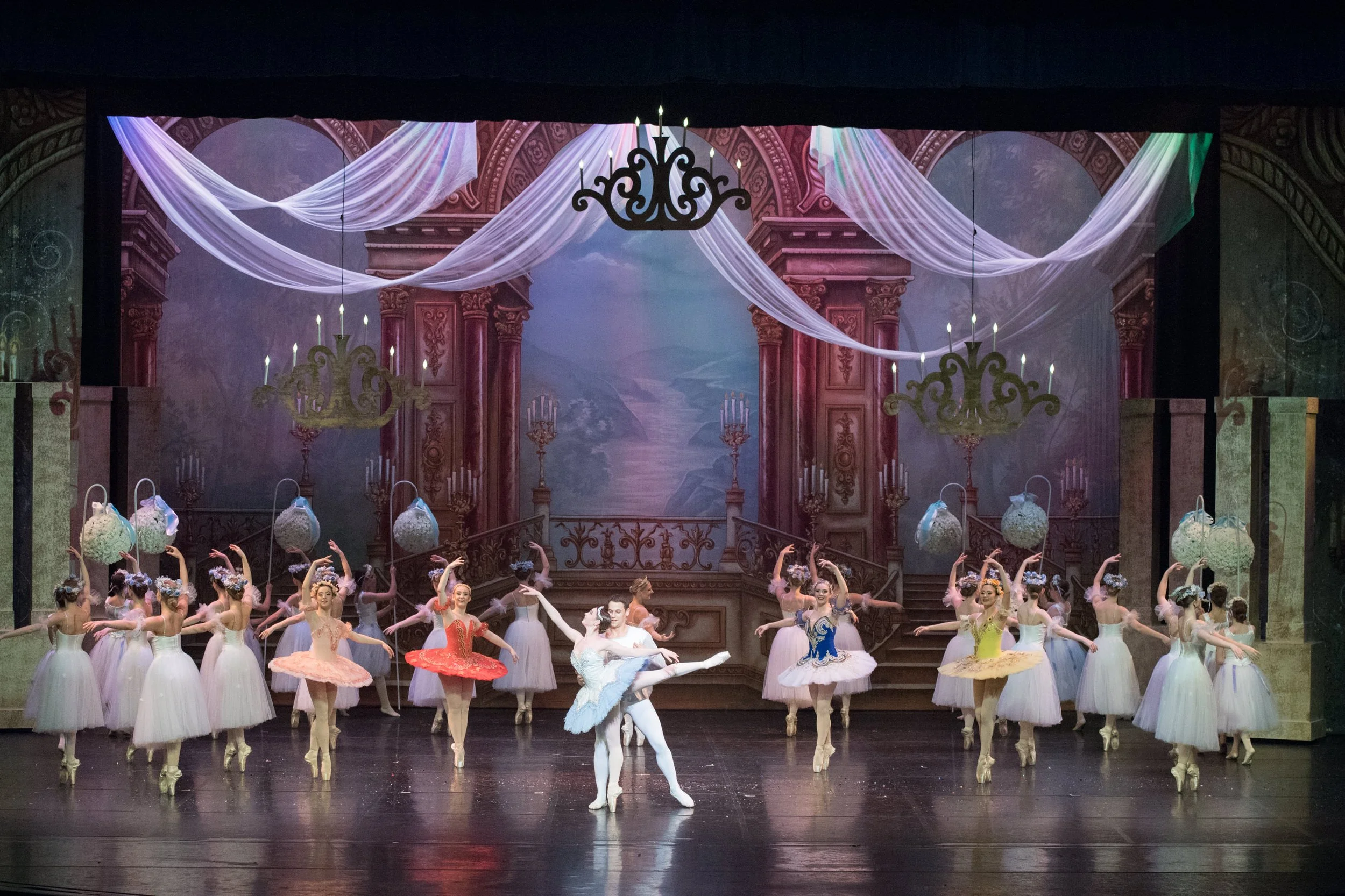 Ballet dancers performing on stage with a backdrop of a grand, ornate theater interior, featuring chandeliers, draped fabric, and a painted landscape. The dancers are in colorful tutus, with some holding flower baskets.