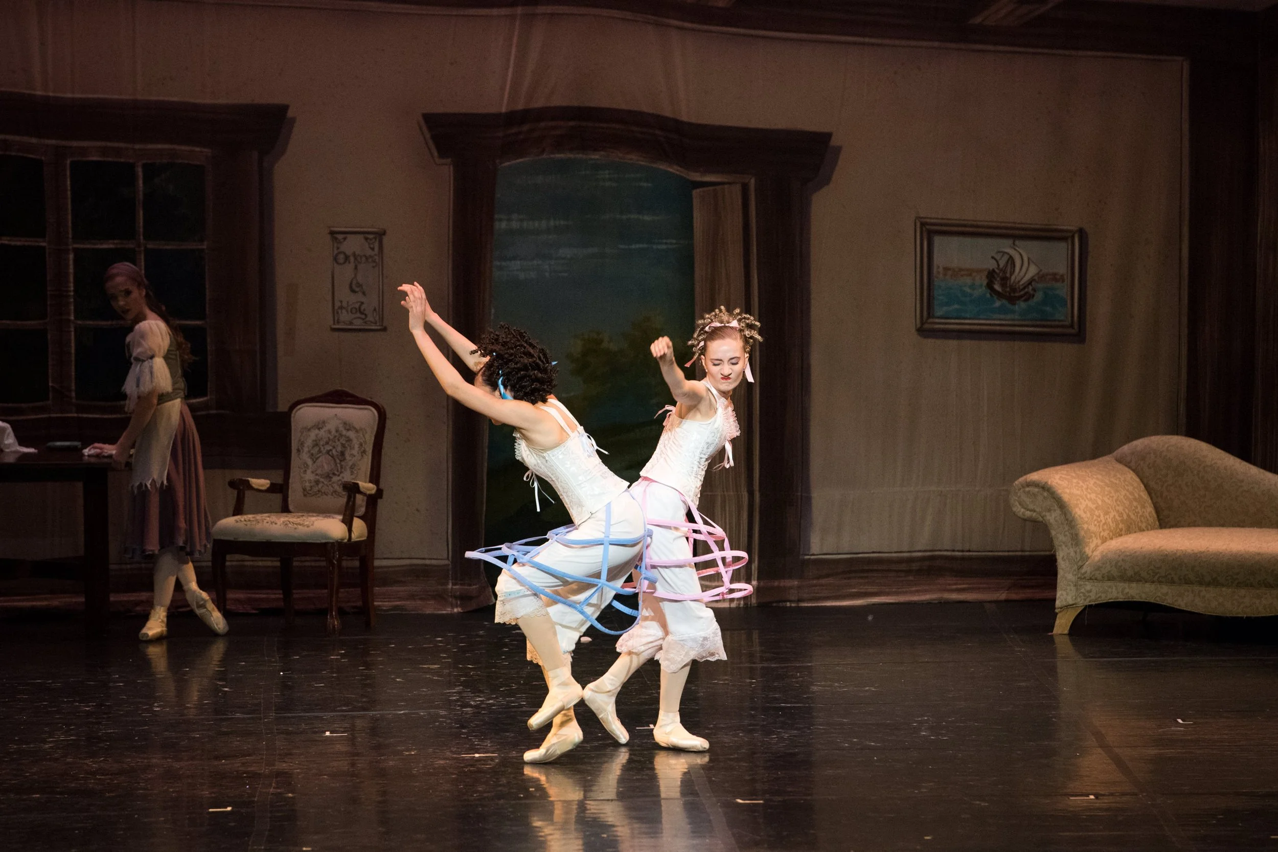 Two ballerinas with colorful hoop skirts performing on stage in front of a painted backdrop, with a third person observing in the background.
