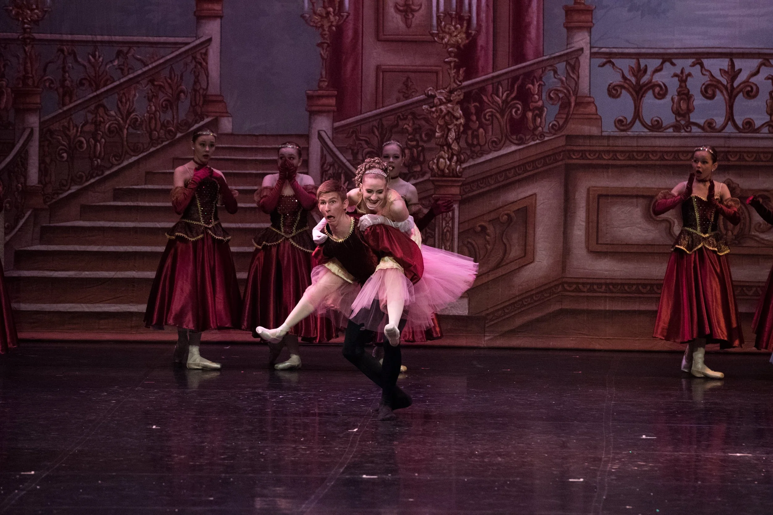 Ballet dancers perform on stage in front of a painted backdrop of a grand staircase and ornate walls, with one dancer lifting another in the foreground.