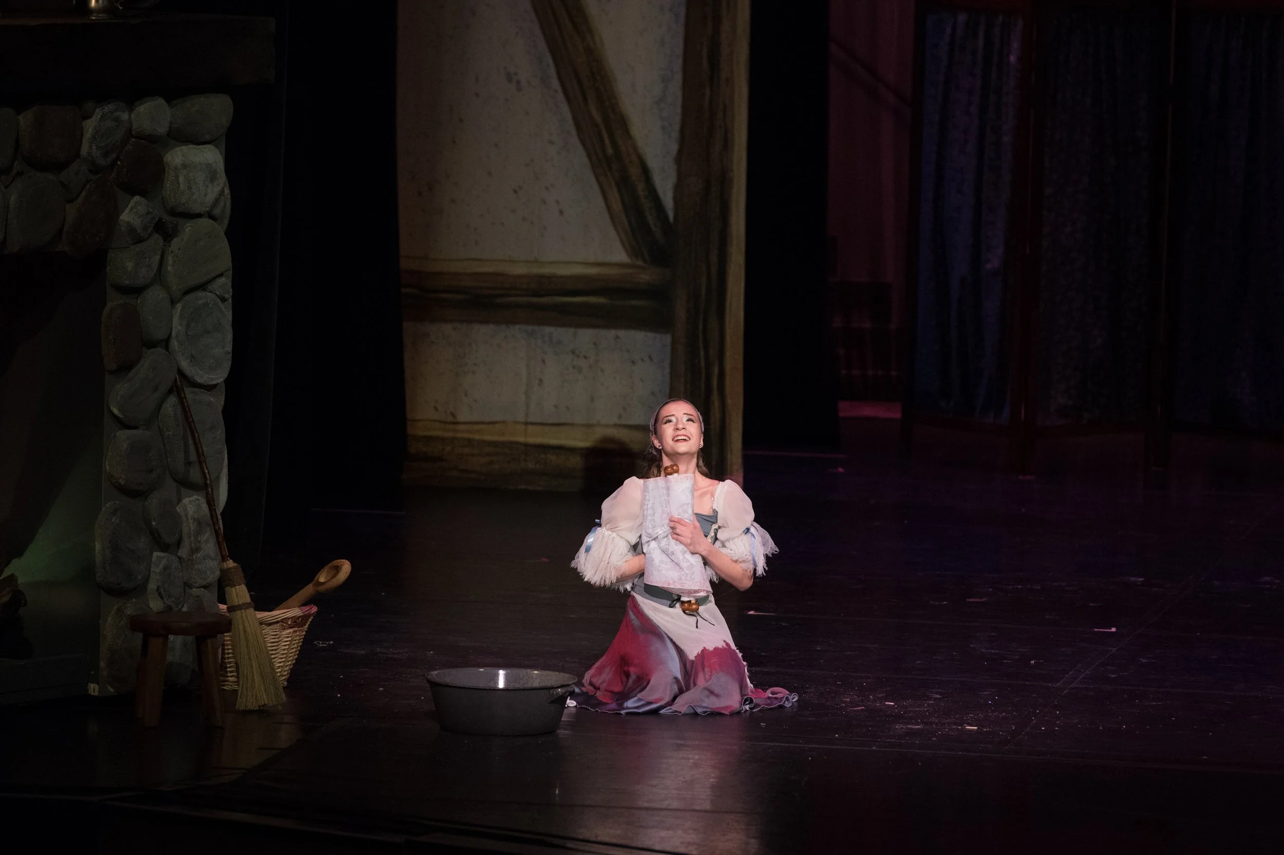A woman dressed in costume kneels on stage, singing or speaking with a joyful expression, surrounded by stage props including a stone-looking wall, a small stool, a broom, a basket, and a basin, with a painted backdrop.