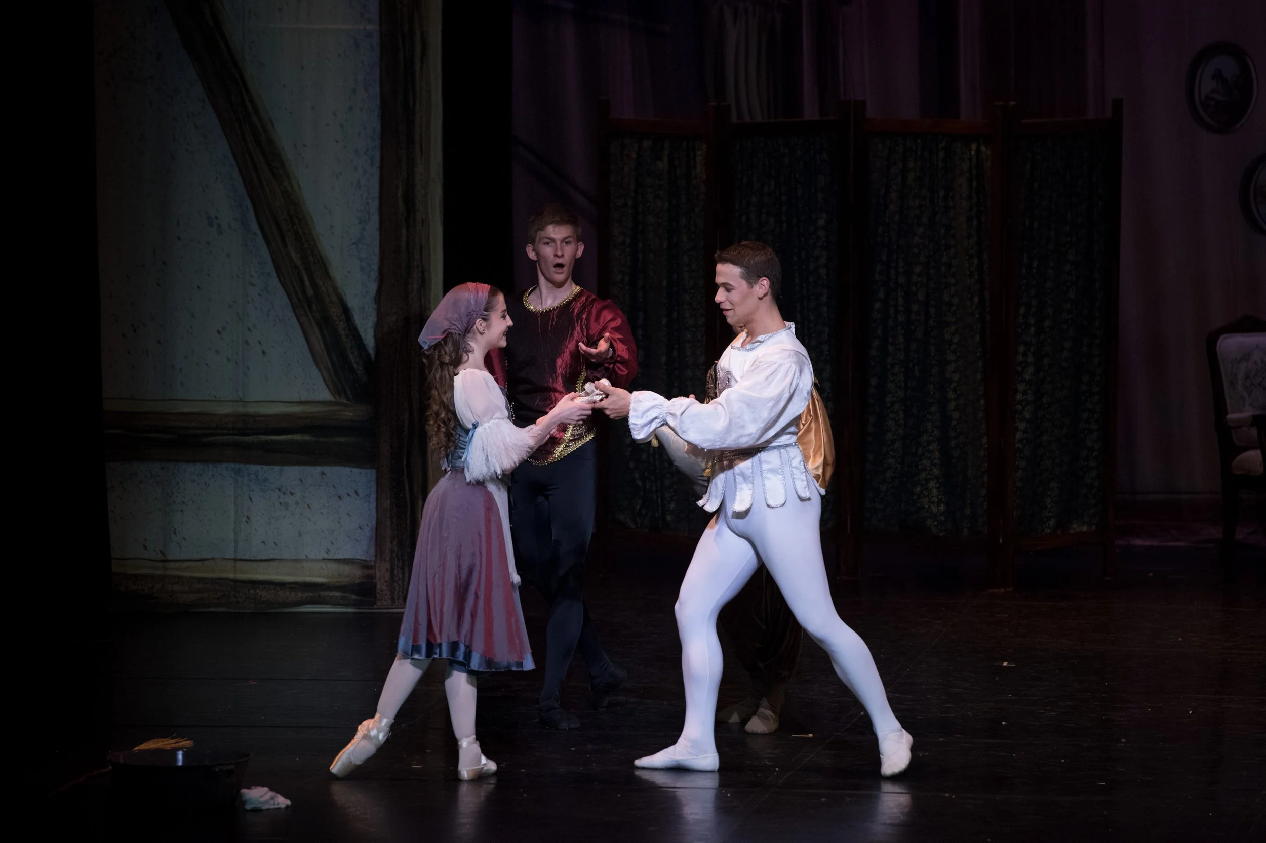 Three ballet dancers in period costumes performing on stage during a play or ballet, with a backdrop of trees and a stage set.