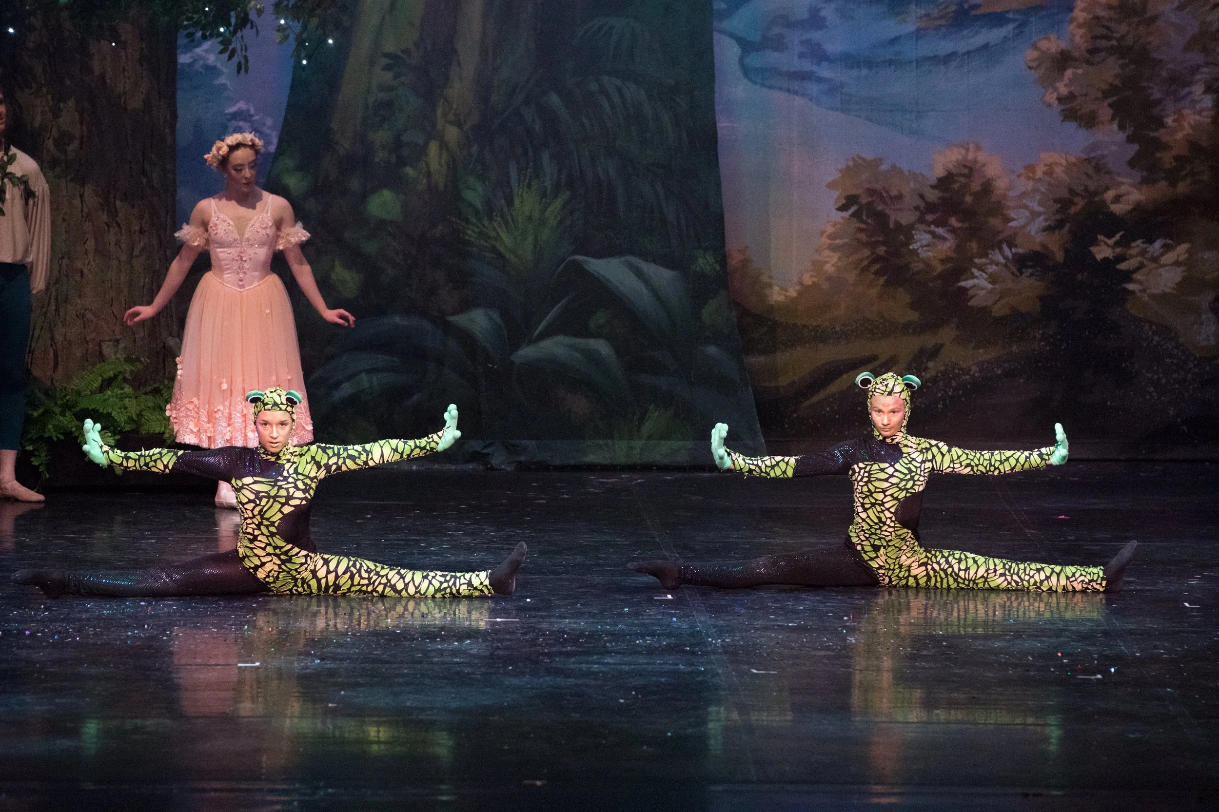 Two performers dressed as frogs in a split pose on stage, with a backdrop of a forest scene and a woman in a pink dress and flower crown watching.
