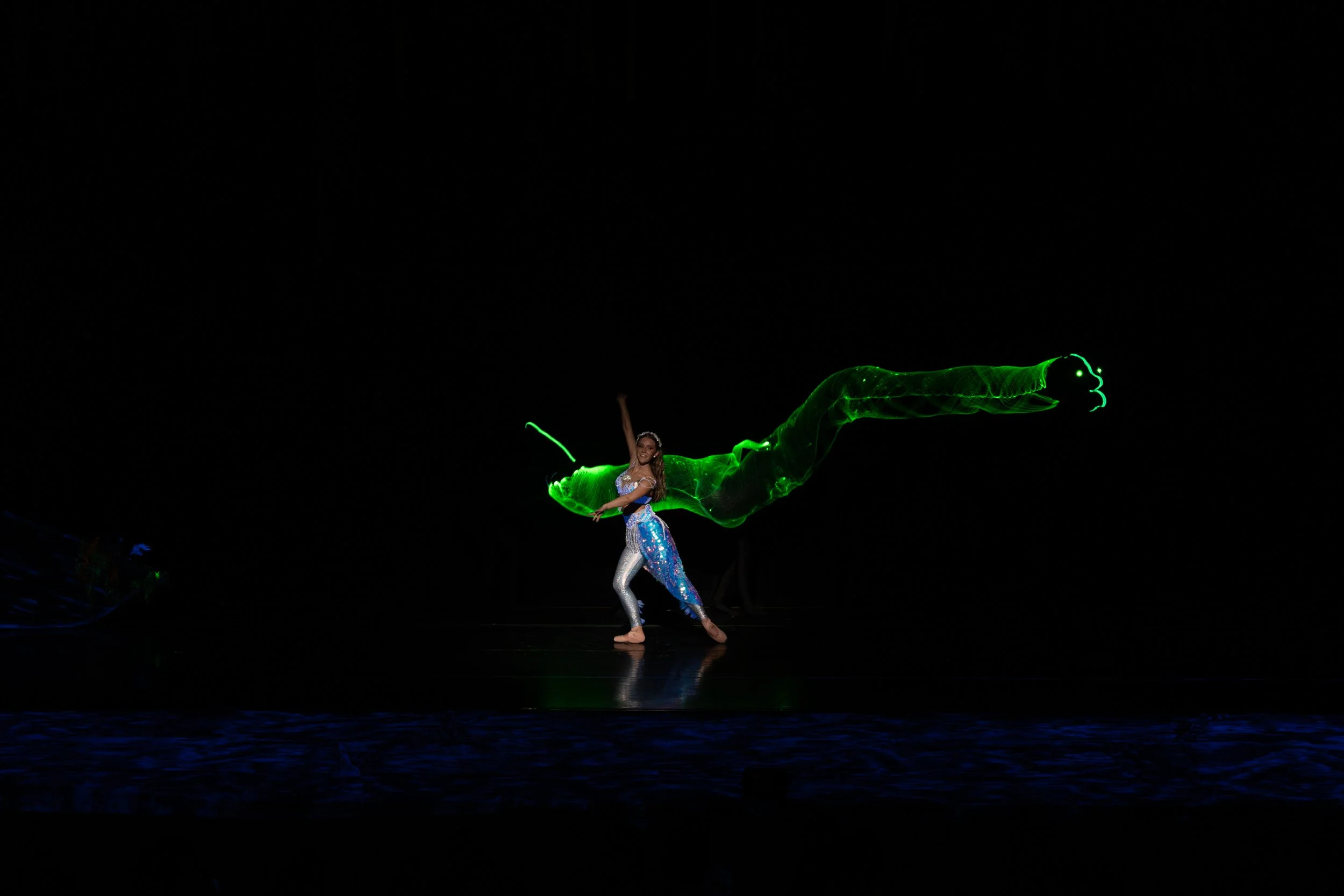 A performer in a shiny costume dances on stage with a green light trail resembling a dragon or serpent in the dark background.