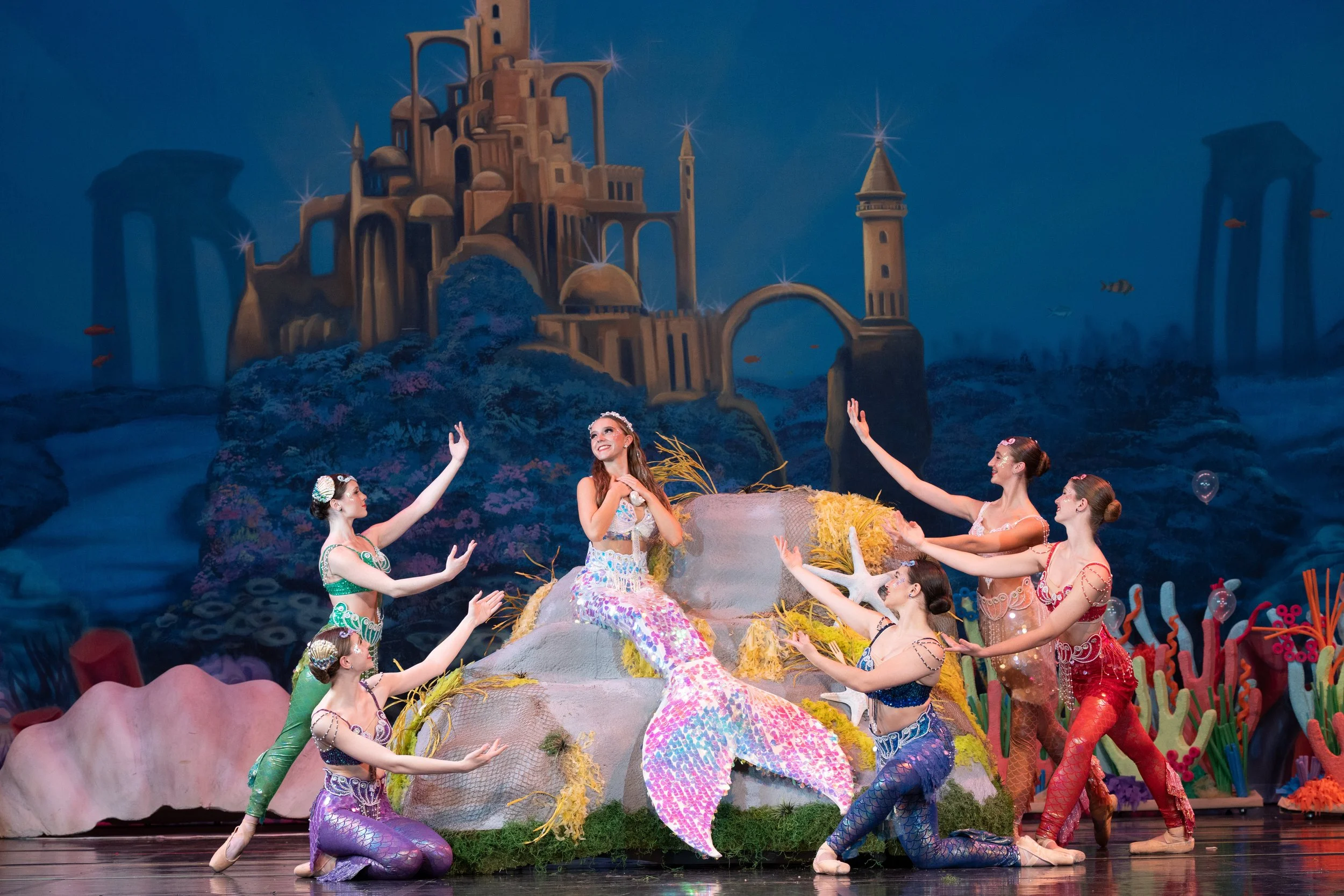 A theatrical dance performance featuring young women dressed as mermaids on stage with an underwater castle backdrop.